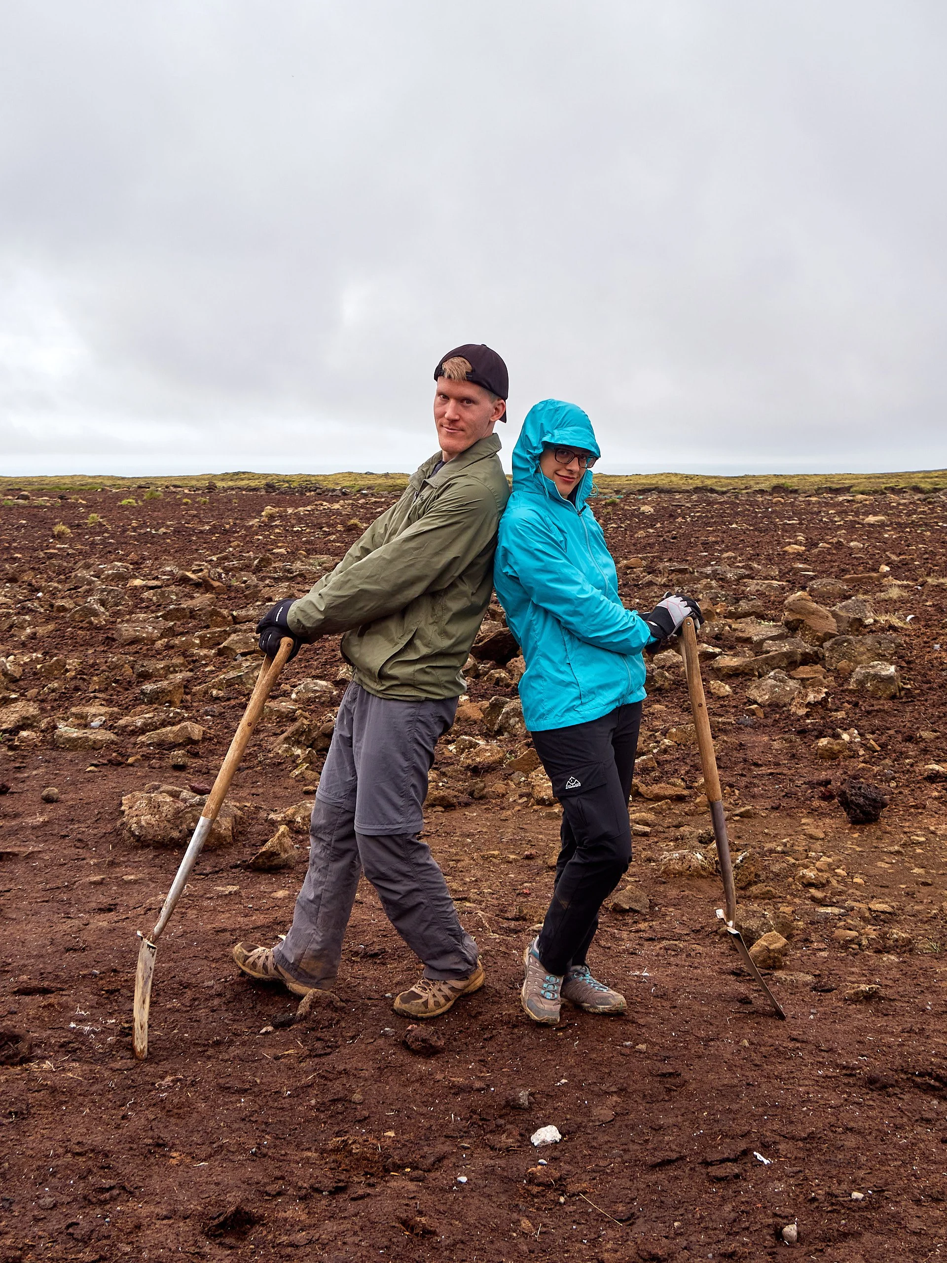 Two people standing back-to-back in a rocky, barren landscape with overcast sky, each holding a hiking pole.