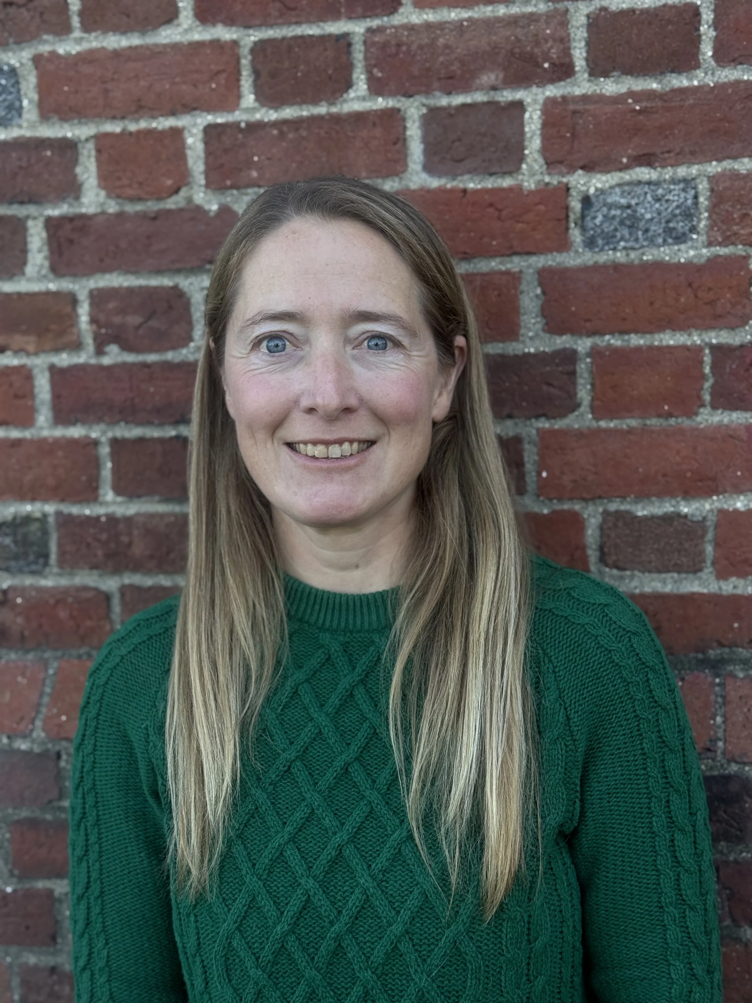 A woman with long blonde hair and blue eyes smiling in front of a red brick wall, wearing a green knitted sweater.