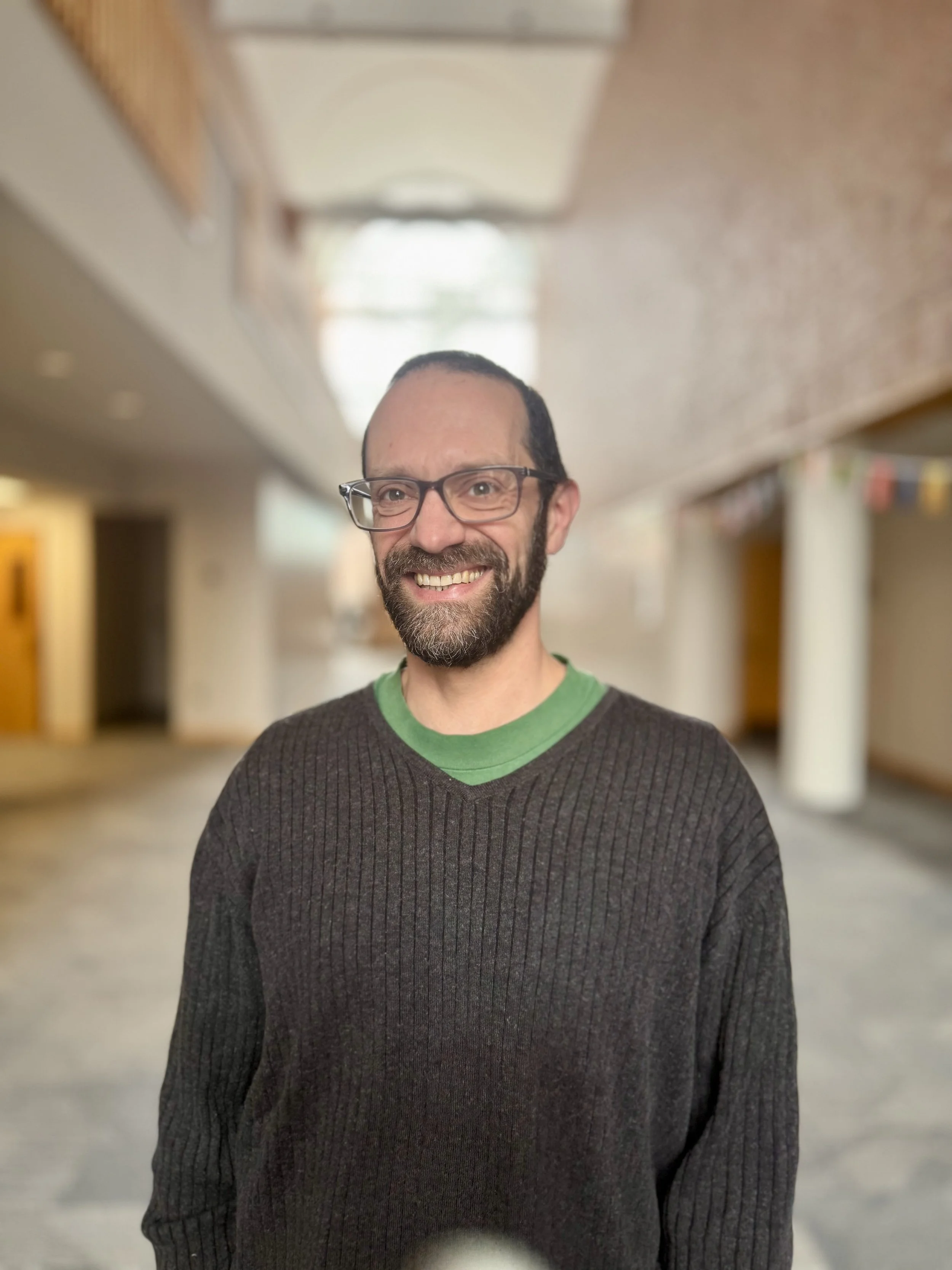 A smiling man with glasses, a beard, wearing a dark gray sweater over a green shirt, standing in a corridor with blurred background.