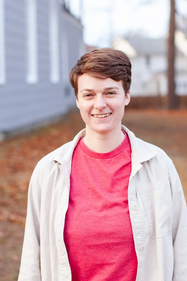 A smiling person with short brown hair, wearing a red shirt and a light-colored jacket, standing outdoors with a blurred background of a house and trees.