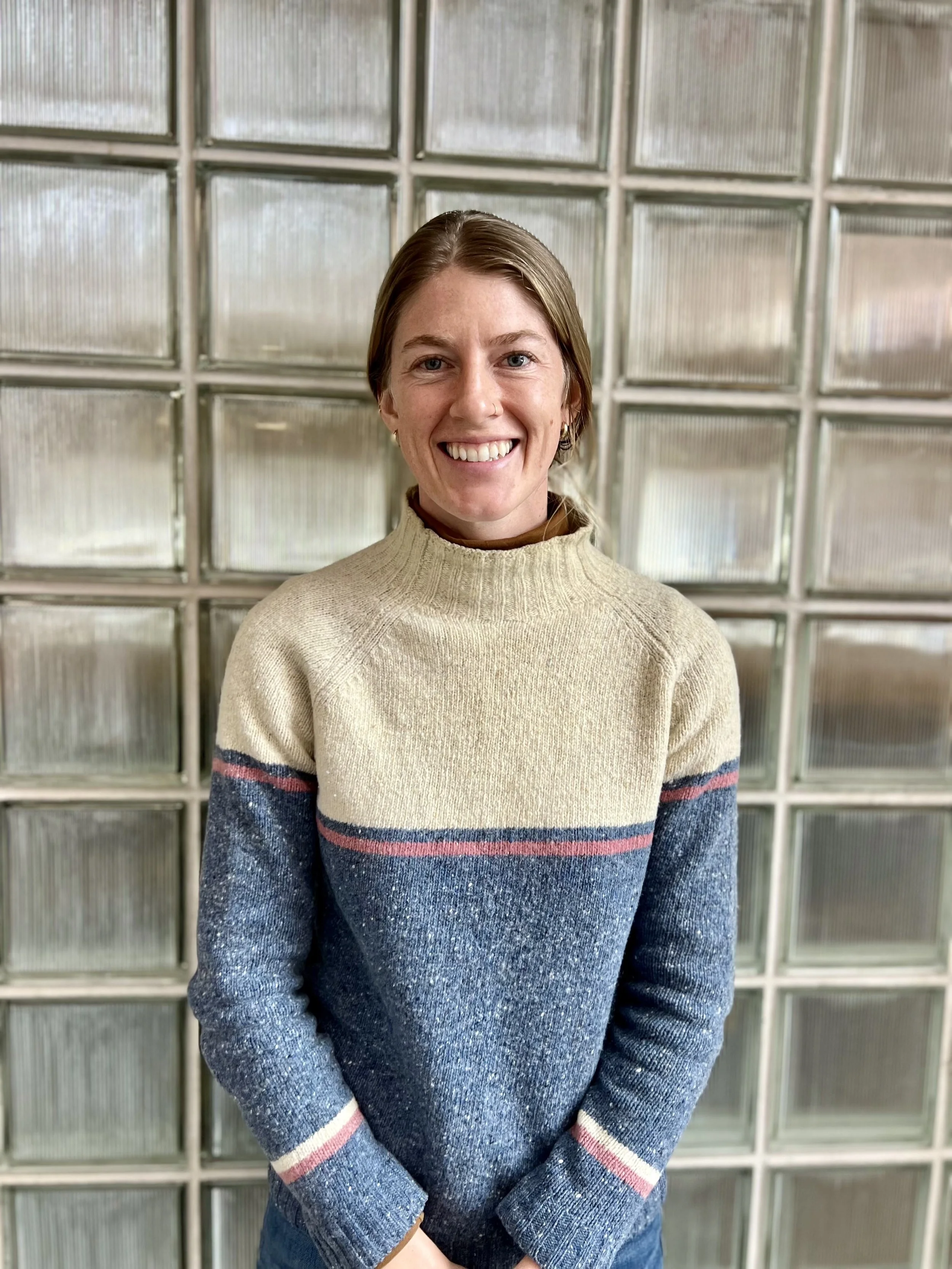 A woman with short brown hair, smiling, wearing a beige sweater with colorful striped sleeves, standing in front of a glass brick wall.