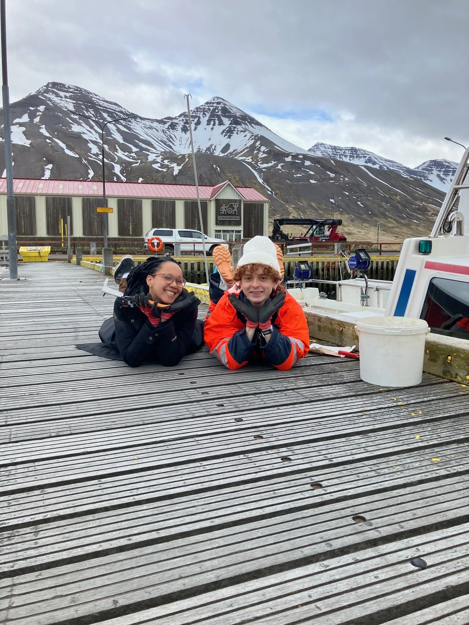Two children lying on a wooden dock near boats with a mountain range in the background.
