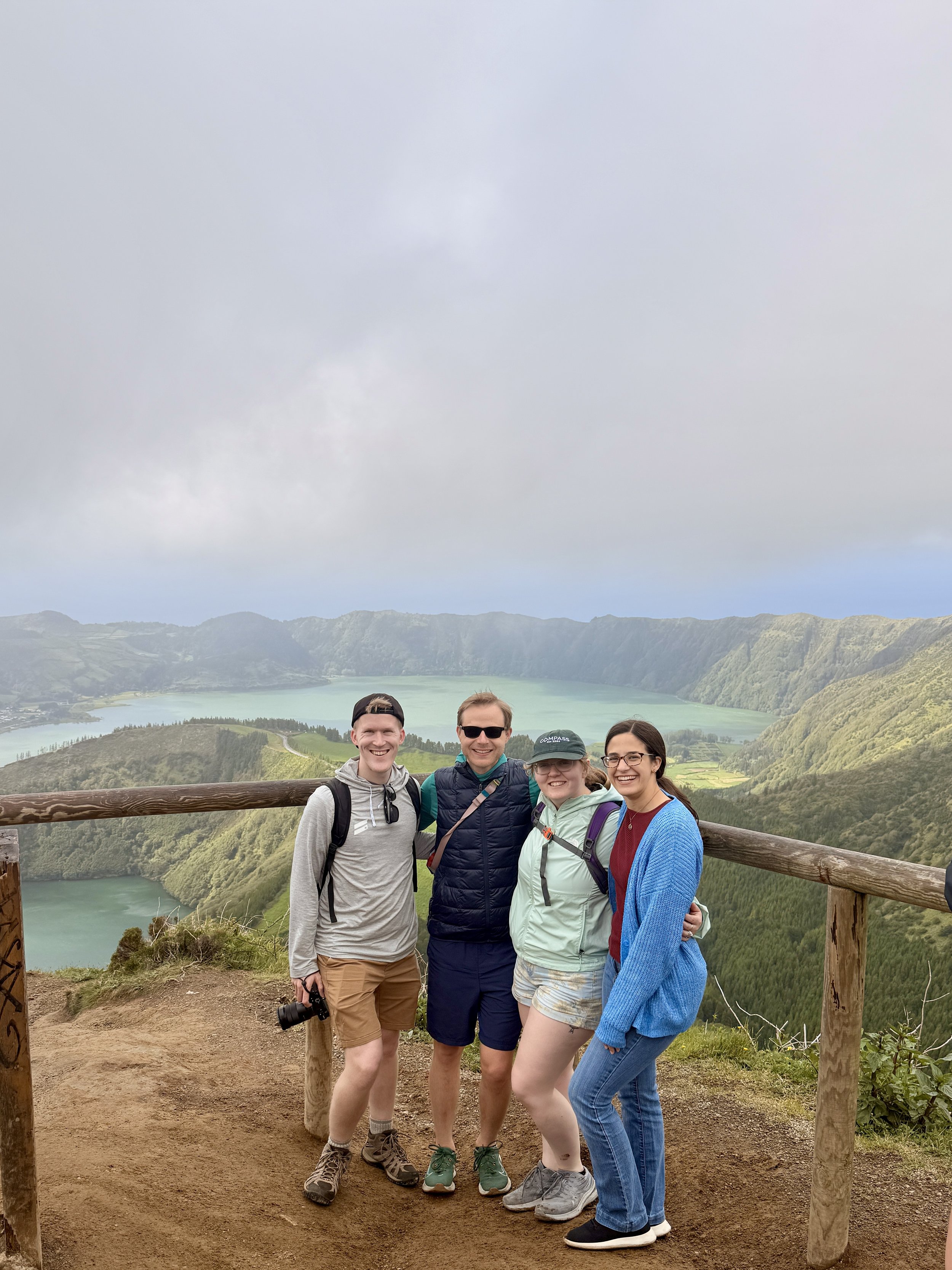 Four people standing together on a hiking trail overlooking a scenic Azores landscape with mountains and a lake. They are smiling and wearing casual outdoor clothing.