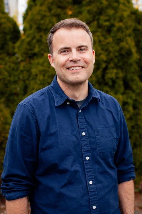 A man smiling outdoors in front of green trees, wearing a navy blue button-up shirt.