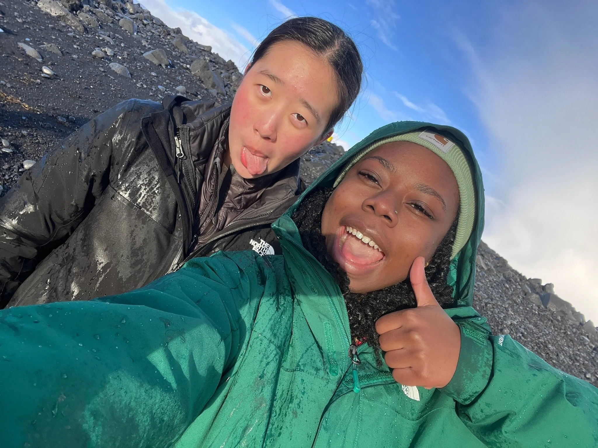 Two smiling individuals sticking out their tongues, wearing waterproof jackets, taking a selfie outdoors on a rocky, volcanic landscape under a partly cloudy sky.