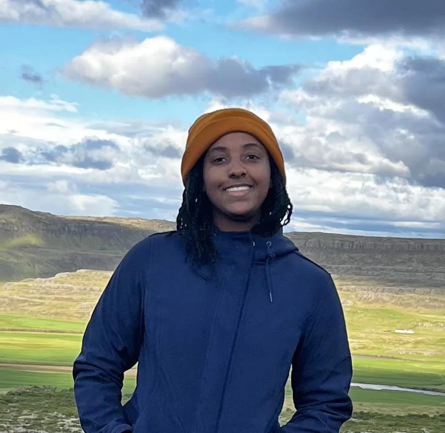 A smiling person with black dreadlocks wearing a mustard-colored beanie and navy blue jacket outdoors with green fields and hillside under a partly cloudy sky.
