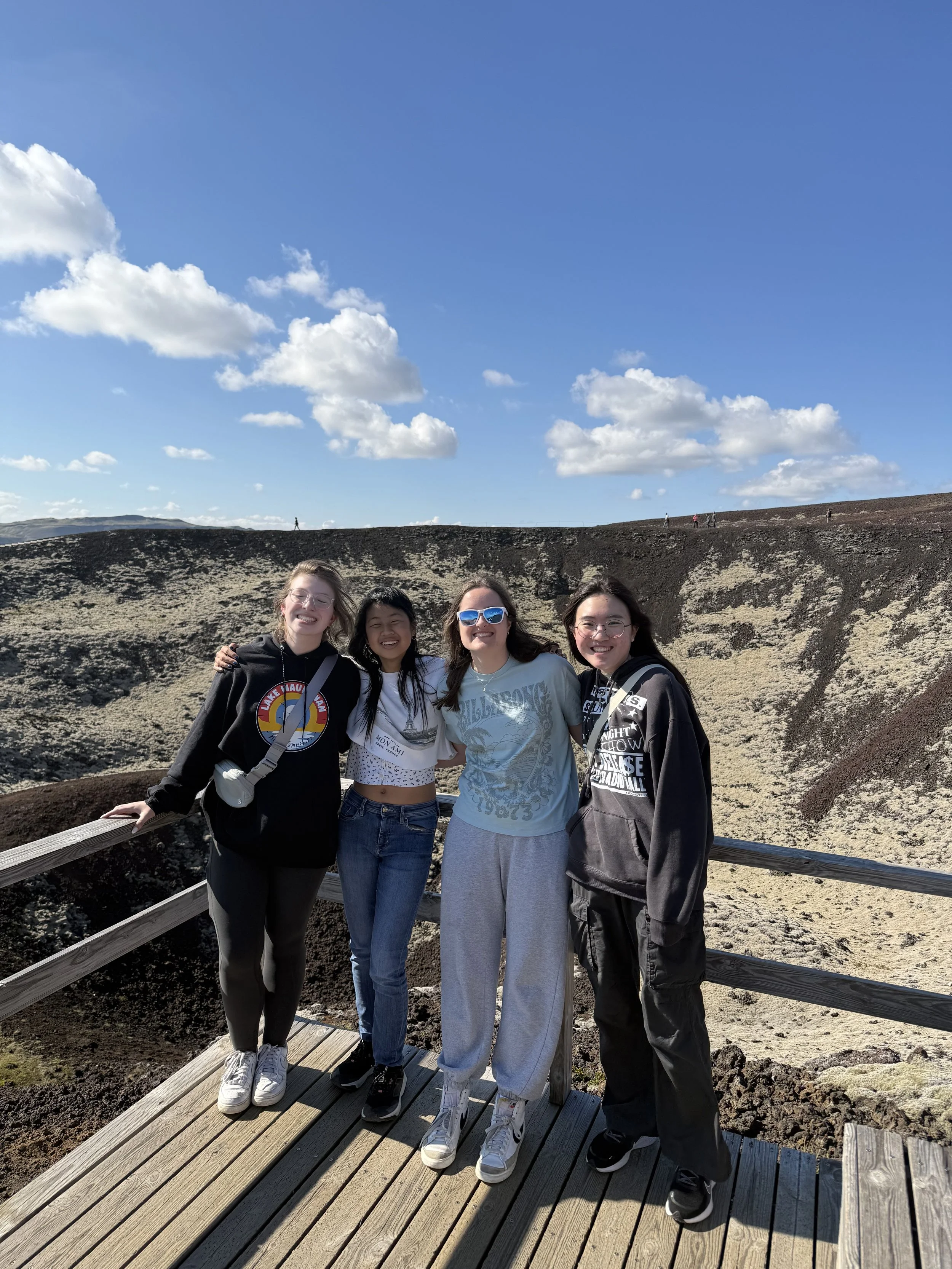 Four women standing together on a wooden viewing platform outdoors with volcanic landscape and blue sky with clouds in the background.