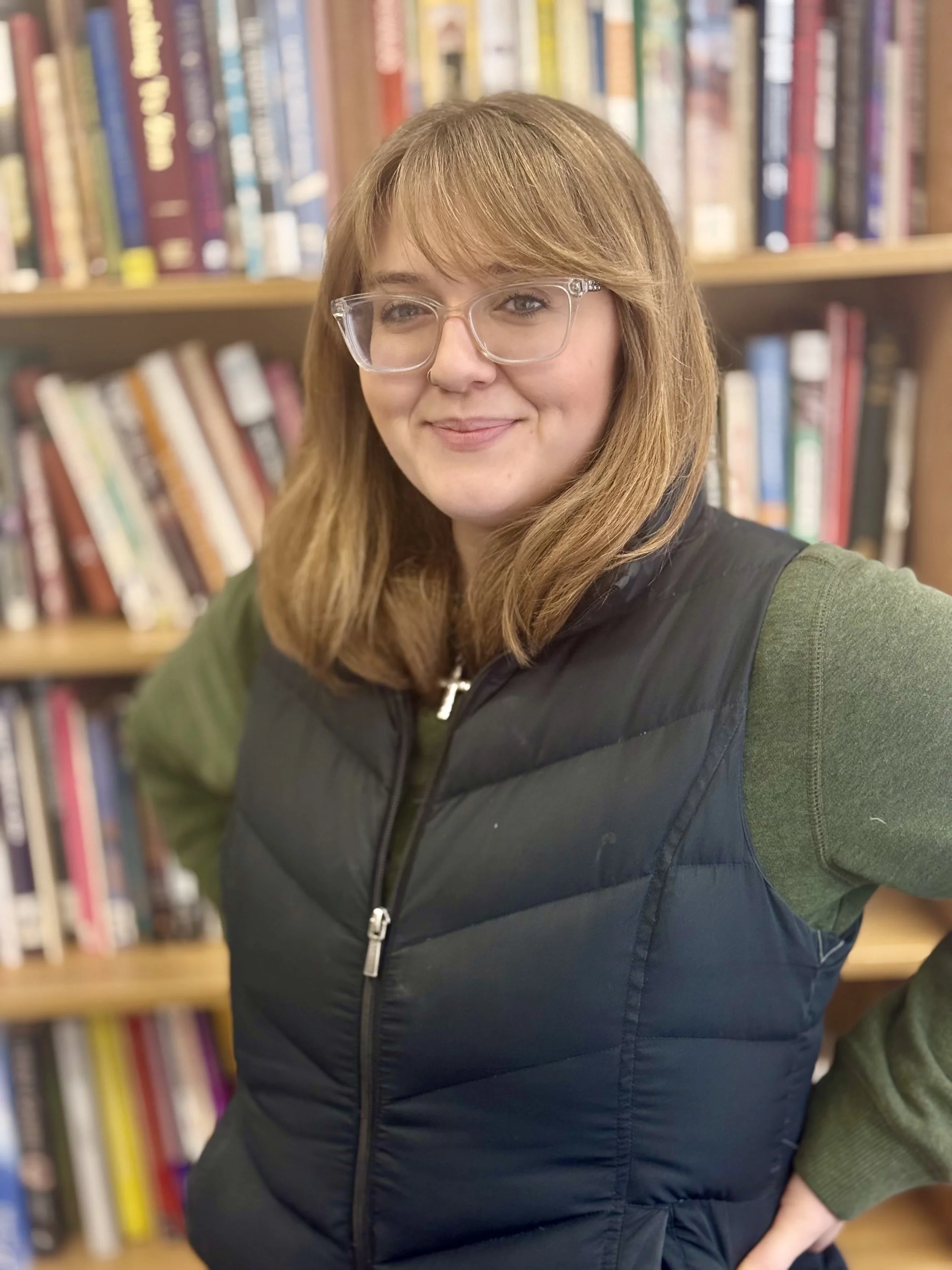 A young woman with red hair wearing glasses and a black puffer vest inside a library, with shelves of colorful books in the background.