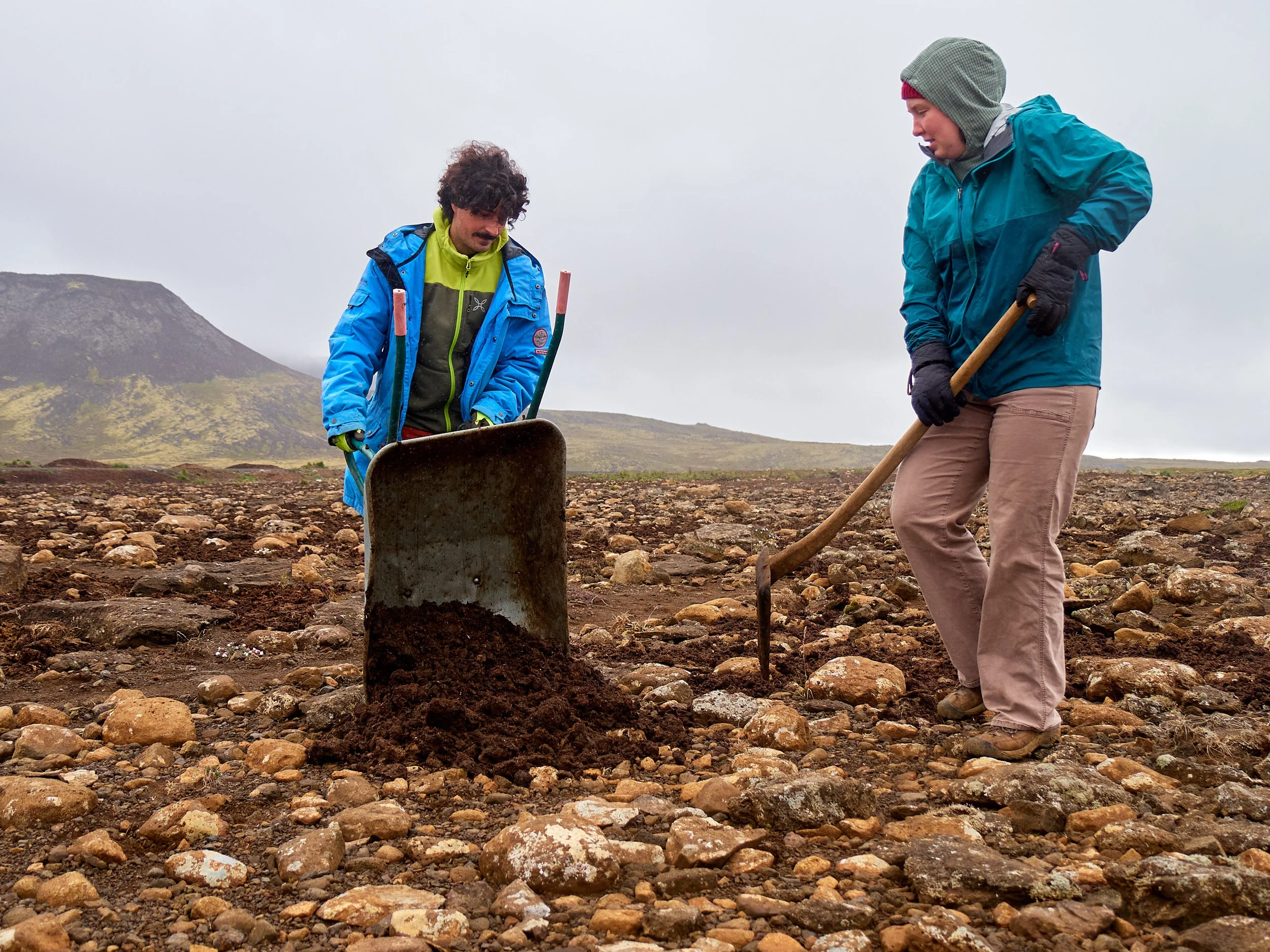 Two men are working outdoors on a rocky terrain, one is digging with a shovel and the other is examining the ground with a wheelbarrow nearby, with mountains and overcast sky in the background.