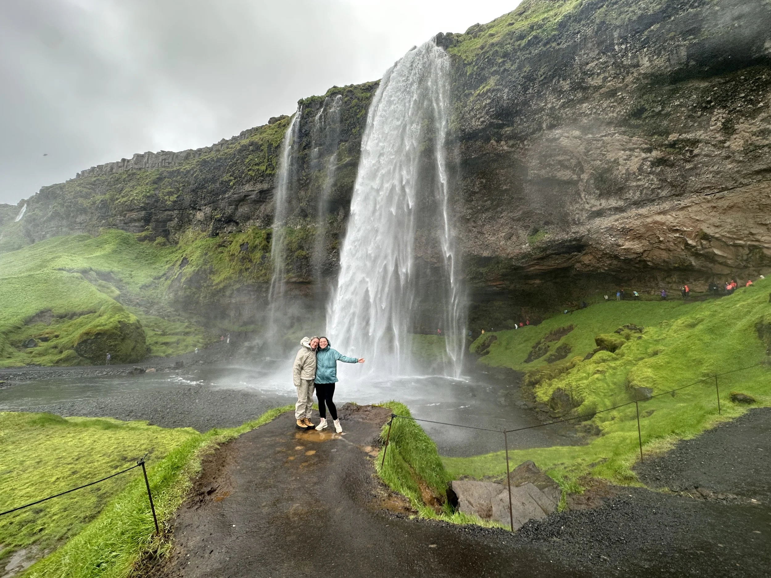 Two people in outdoor gear standing together in front of a large waterfall with a rocky cliff and lush green surroundings, overcast sky.