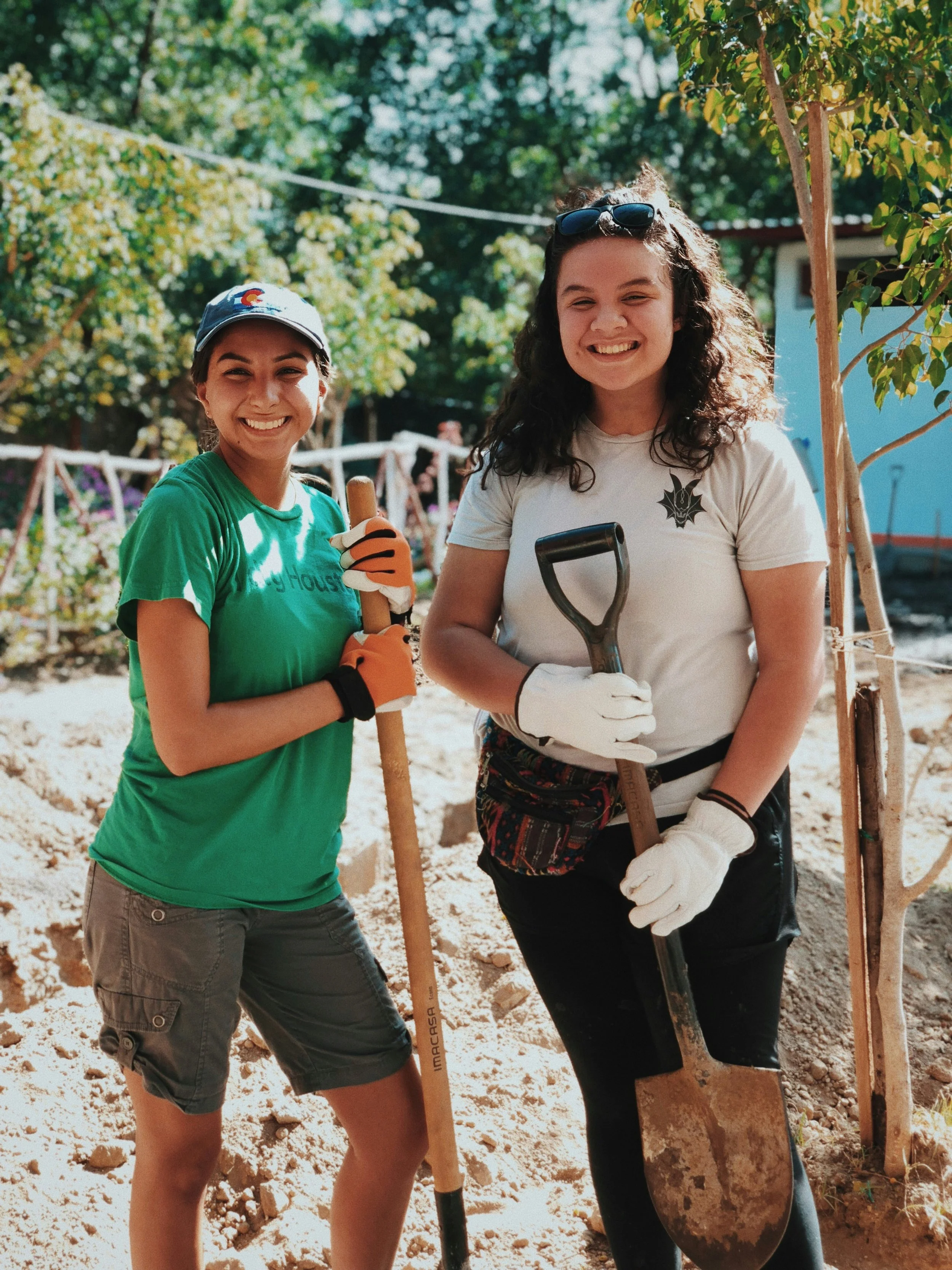 Two young women smiling outdoors during a gardening or landscaping activity, holding shovels, with trees and a building in the background.