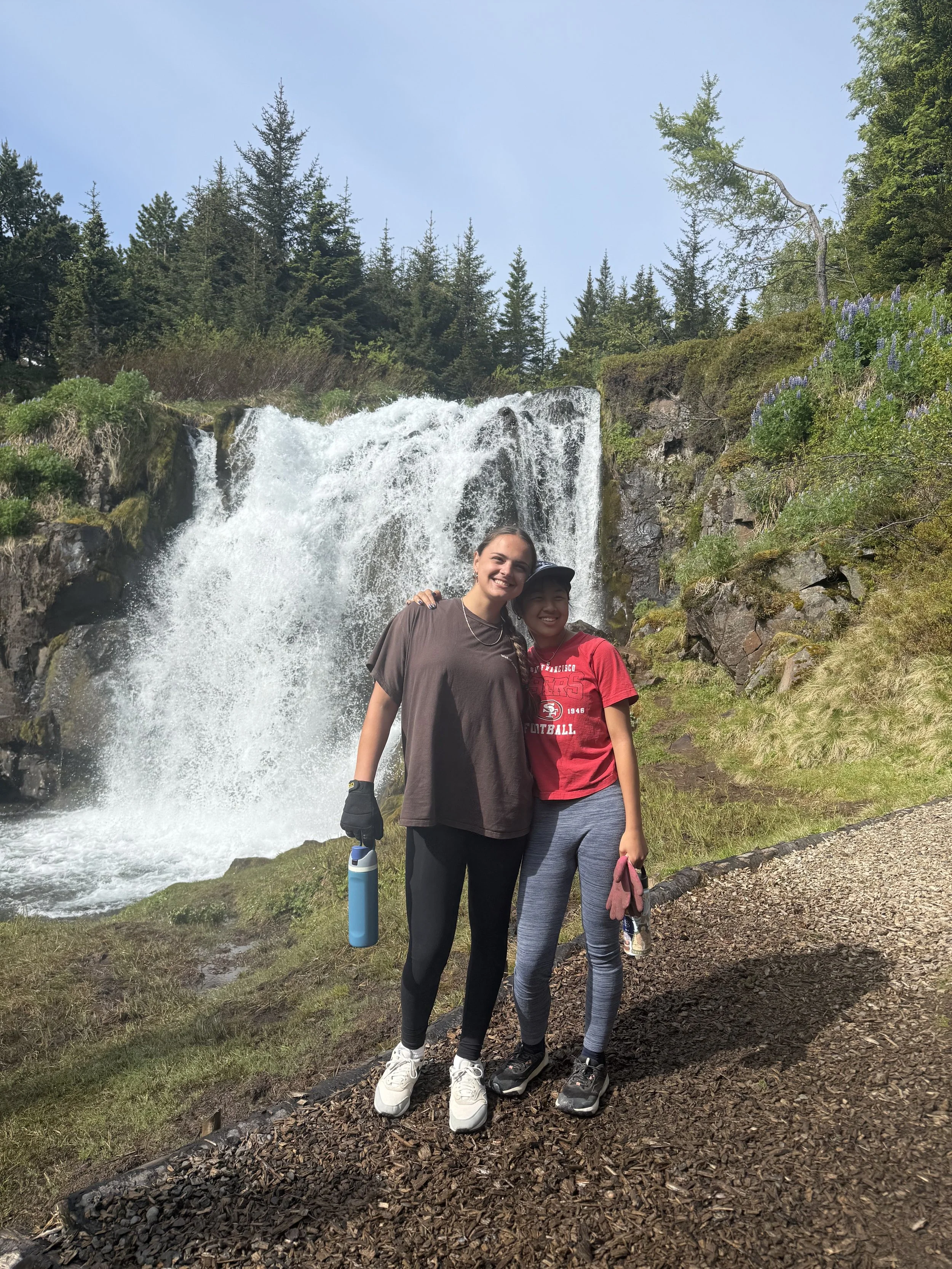 Two women standing in front of a waterfall in a forest setting, smiling, with trees and plants surrounding them.