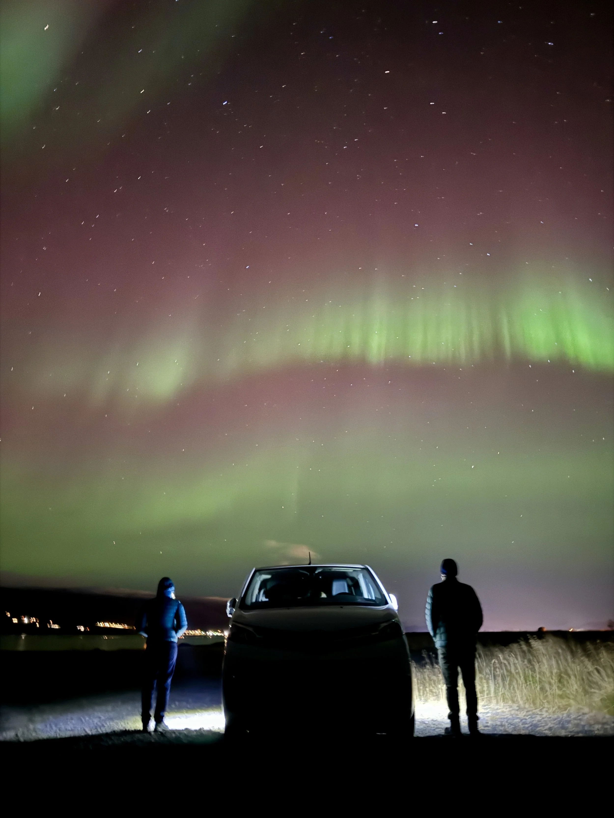 People standing in front of a car watching the Northern Lights over the night sky.