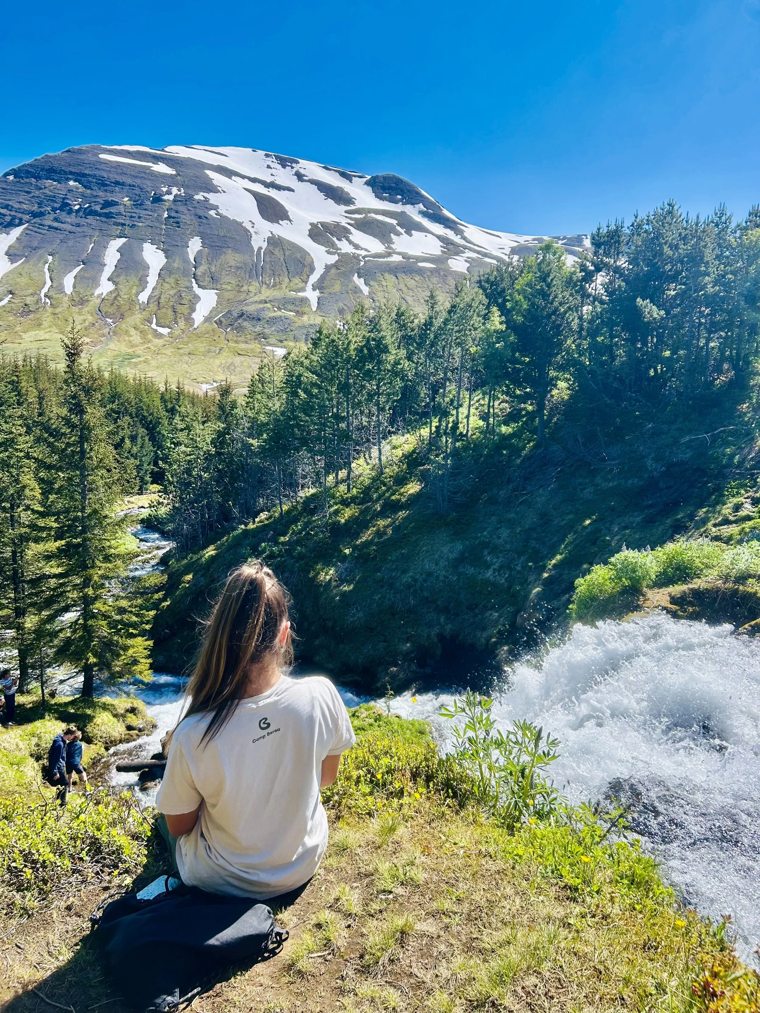 A young woman with brown hair tied back, wearing a white t-shirt, kneeling on grassy ground near a river, with a forest and a snow-capped mountain in the background under a bright blue sky.
