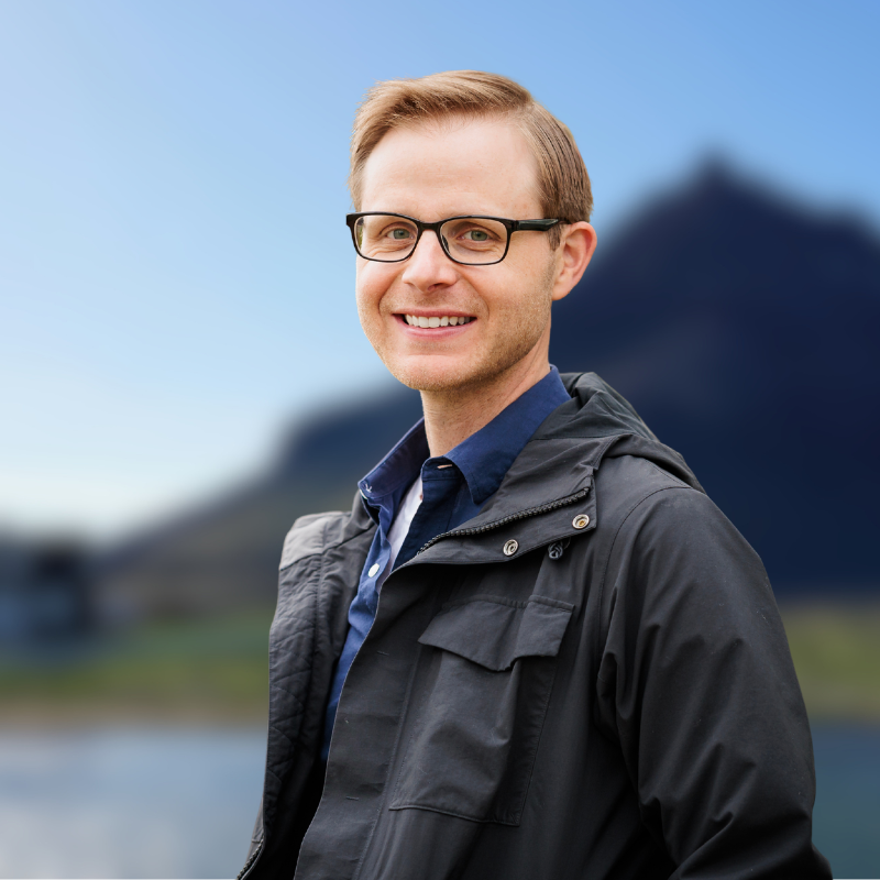 A smiling man with glasses, wearing a blue shirt and a black jacket, standing outdoors with a mountain and blue sky in the background.