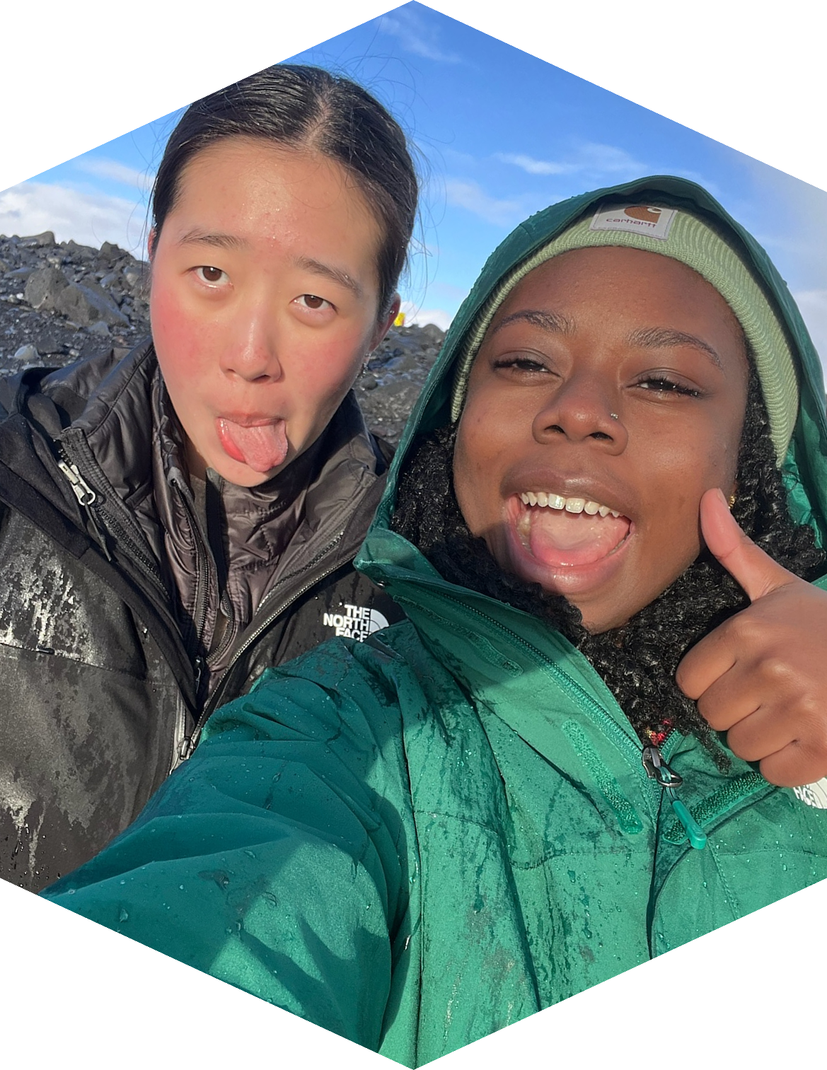Two women outdoors taking a selfie, one is making a playful face with her tongue out, and the other is smiling with her mouth open and thumb up, wearing outdoor jackets and beanie in a rocky landscape.