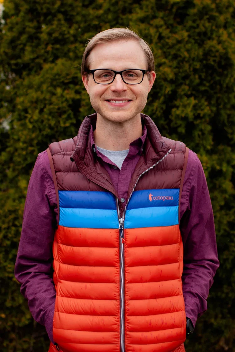 A young man with glasses, short light brown hair, wearing a purple shirt and a colorful puffy vest with blue, red, and purple sections, standing outdoors with green foliage in the background.
