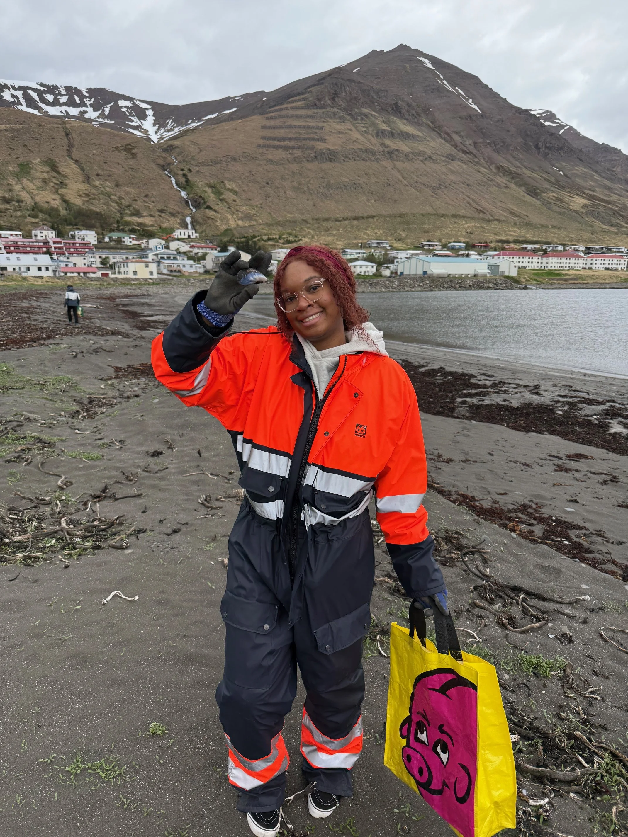 A woman with curly red hair, wearing glasses, an orange and black jacket, black pants, and black shoes, holding up a small fish in her gloved hand and smiling at the camera on a beach with a mountainous background and a town at the base of the mountain.