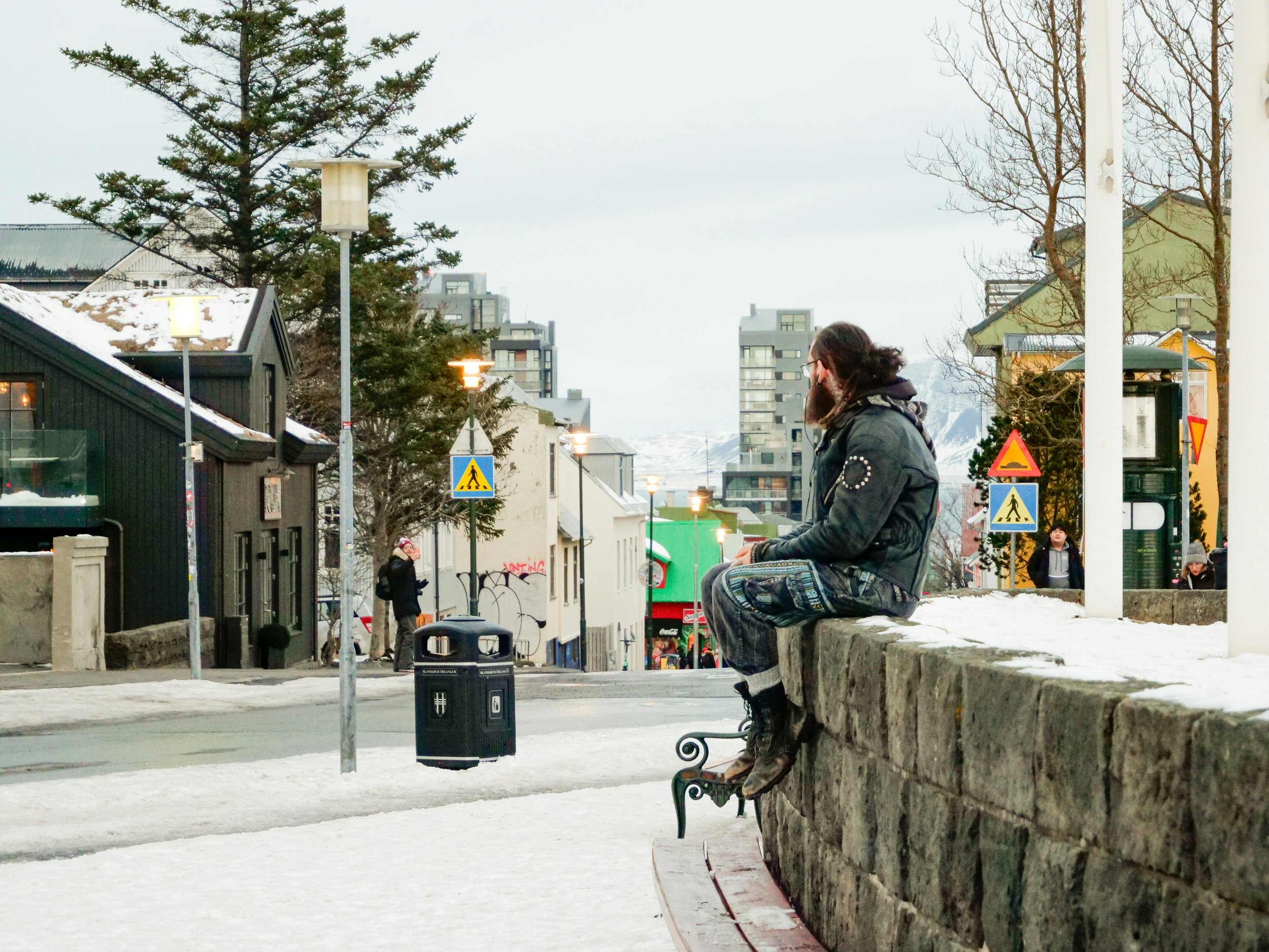 A person sitting on a stone wall in a snowy outdoor urban setting, wearing a black jacket, with other people walking in the background, and a mix of modern buildings and trees around.
