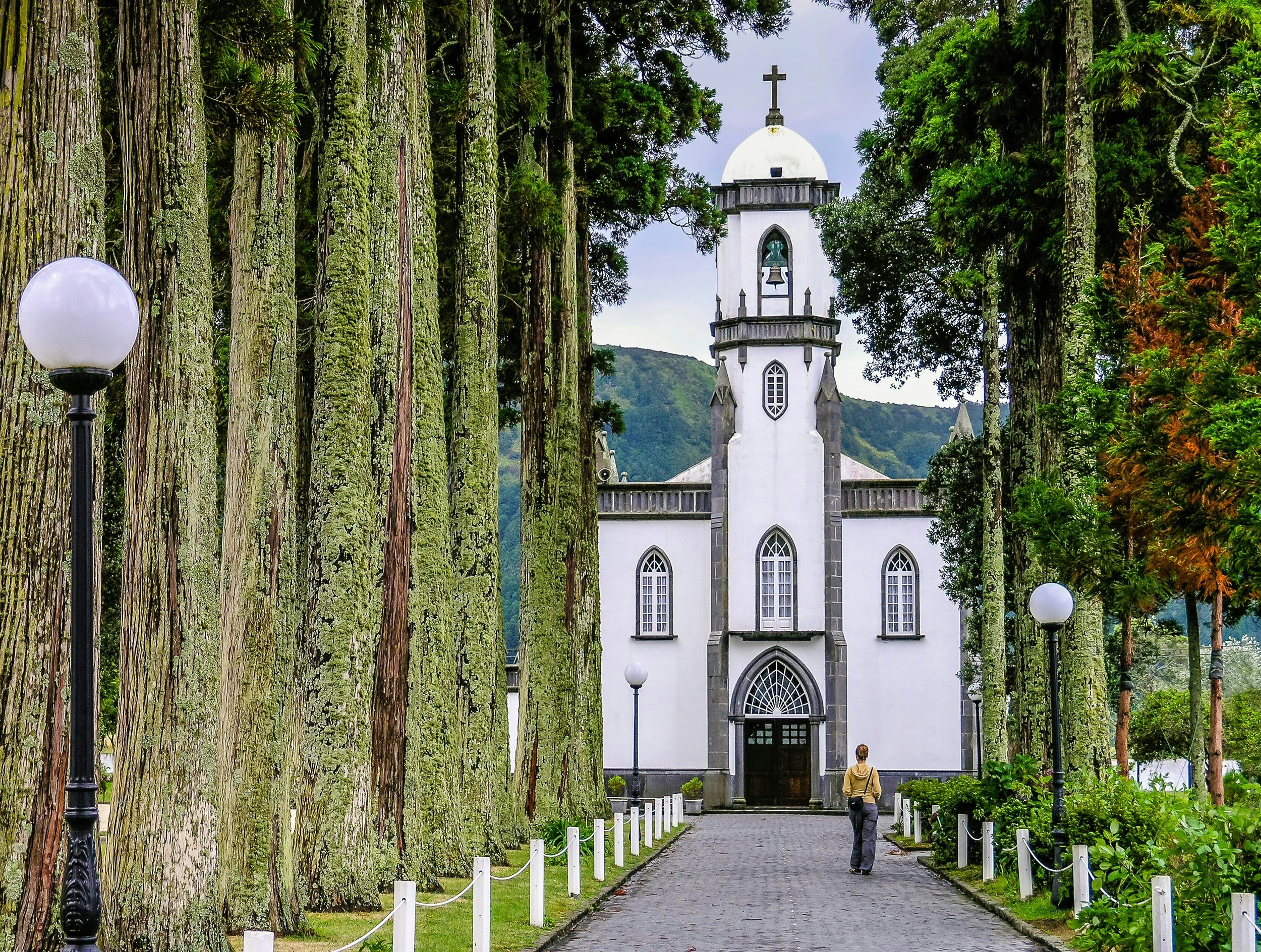 A pathway lined with trees and lampposts leads to a white church with a cross on top, set against a backdrop of green hills and cloudy sky.