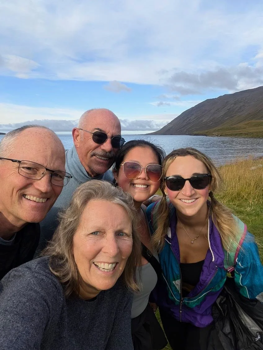 Group of five people taking a selfie outdoors with a lake, mountains, and cloudy sky in the background.
