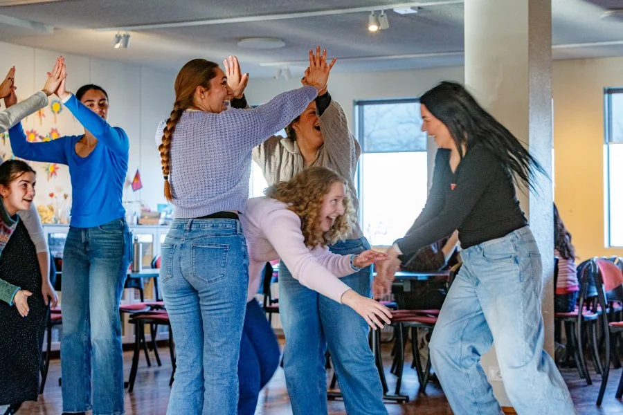 Group of young women and girls laughing and playing together indoors, some raising their hands while others join in a playful dance or game, with chairs and windows in the background.