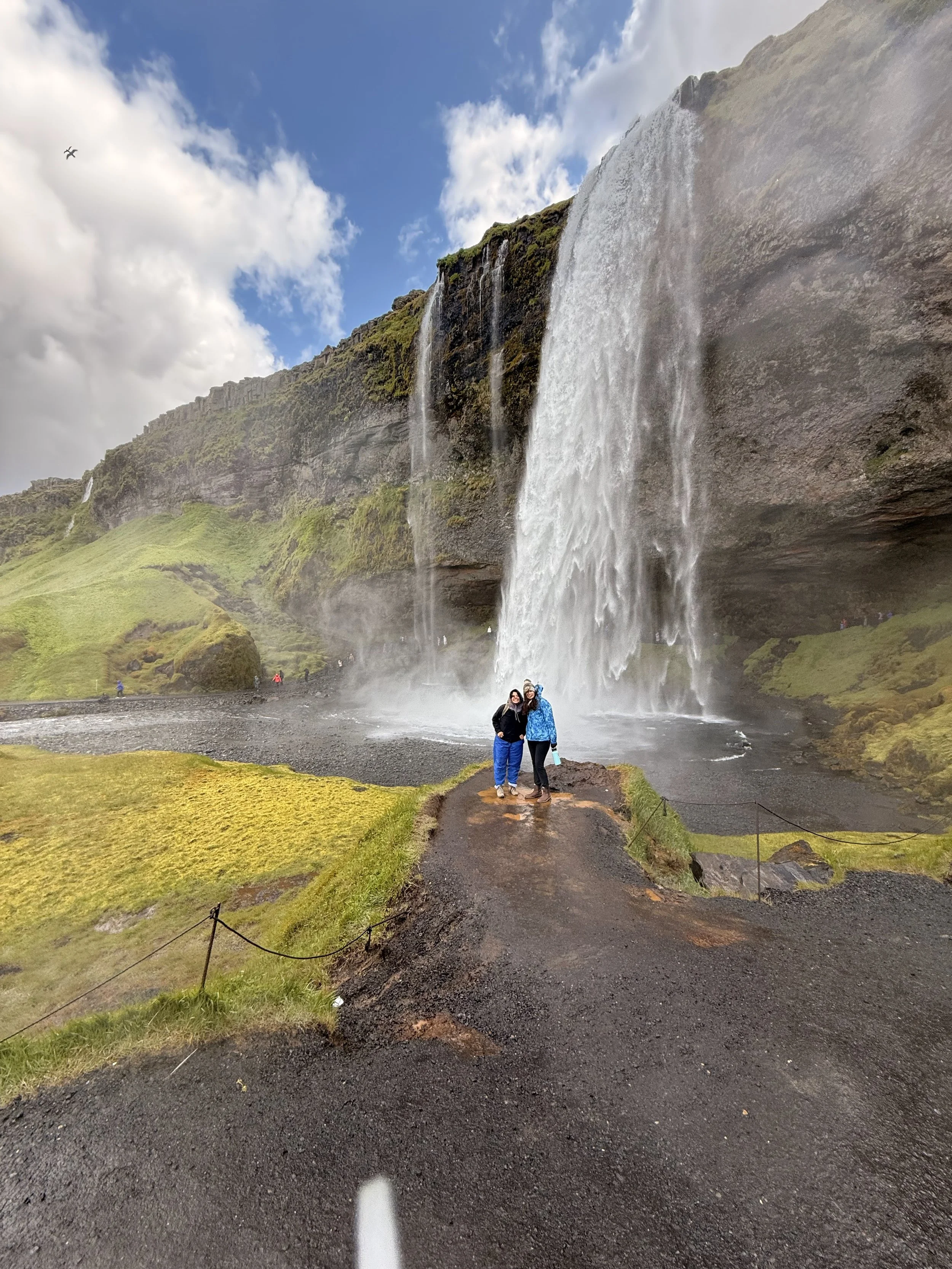 Two people standing on a muddy path in front of a large waterfall with mist rising from the base, surrounded by green moss-covered rocks and grass, with a partly cloudy sky overhead.