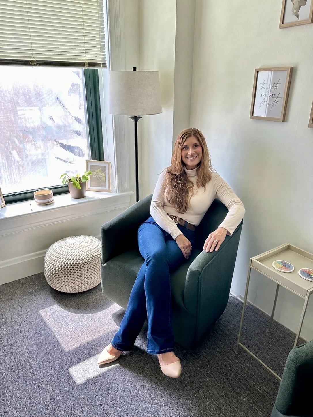 A woman with long, wavy hair sitting happily in a green armchair in a well-lit room with a large window, decorated with wall art and small decorative items on a side table and windowsill.