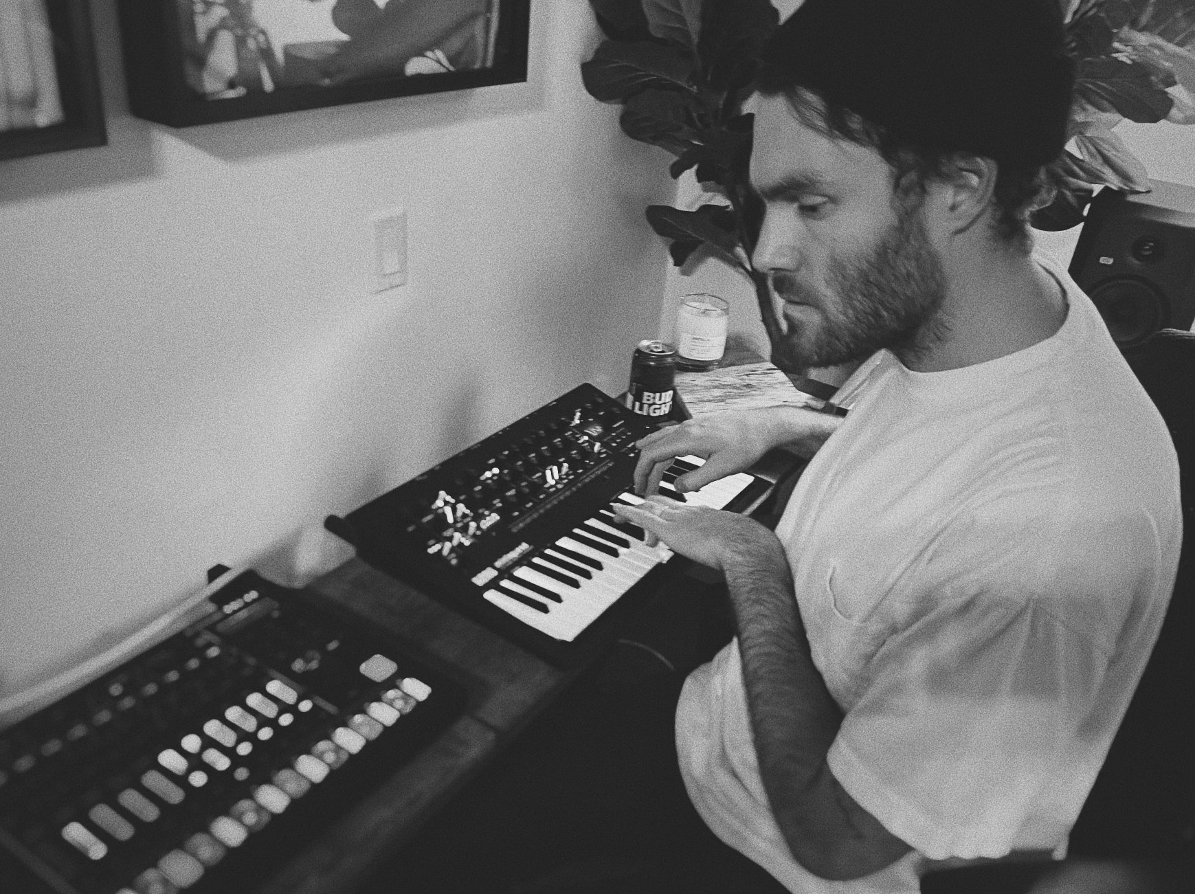 A man with curly hair and a beard playing a small keyboard synth in a room, with framed art on the wall, a large leafy plant, and a speaker in the background.
