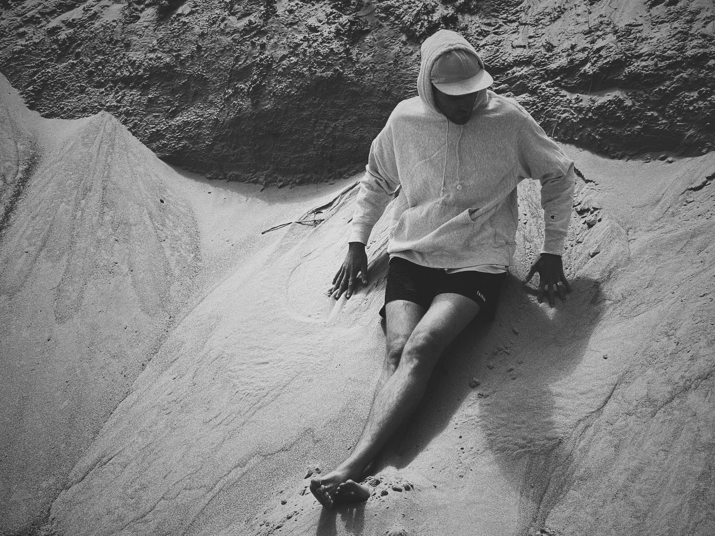 A person sitting on sand dunes, wearing a hoodie, shorts, and a cap, with one hand on the sand and the other resting on their knee, surrounded by textured sand and a dark, rugged background.