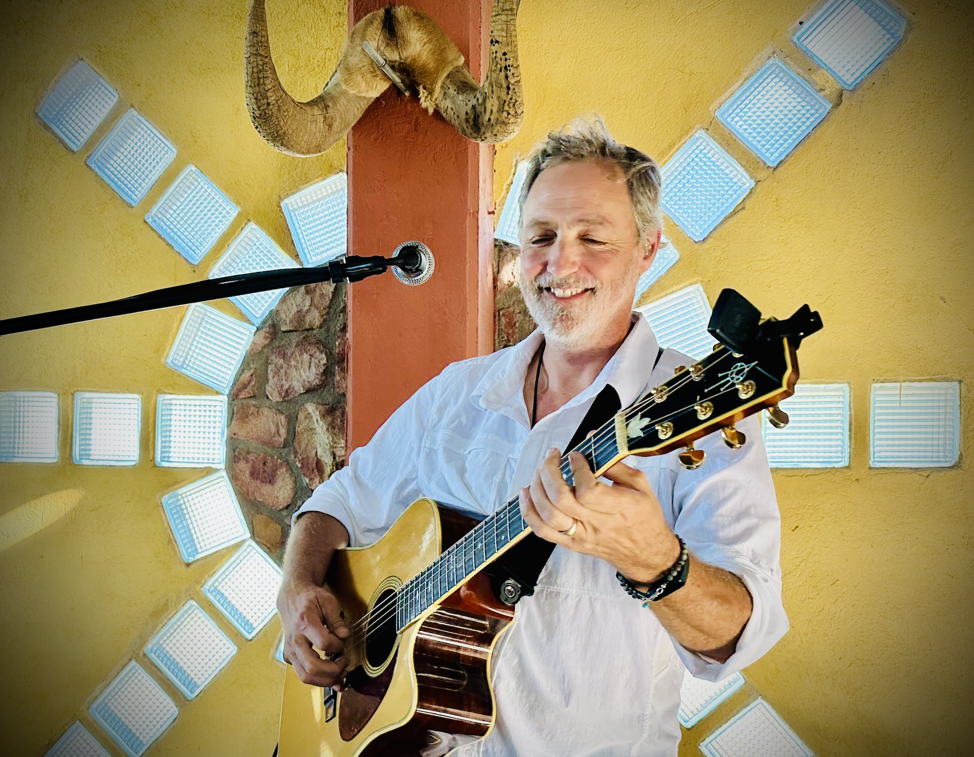 A smiling man with gray hair and a beard playing an acoustic guitar indoors. He is wearing a white shirt and black bracelets, standing in front of a yellow wall with decorative glass block windows and a stone column. There is a microphone in front of him.