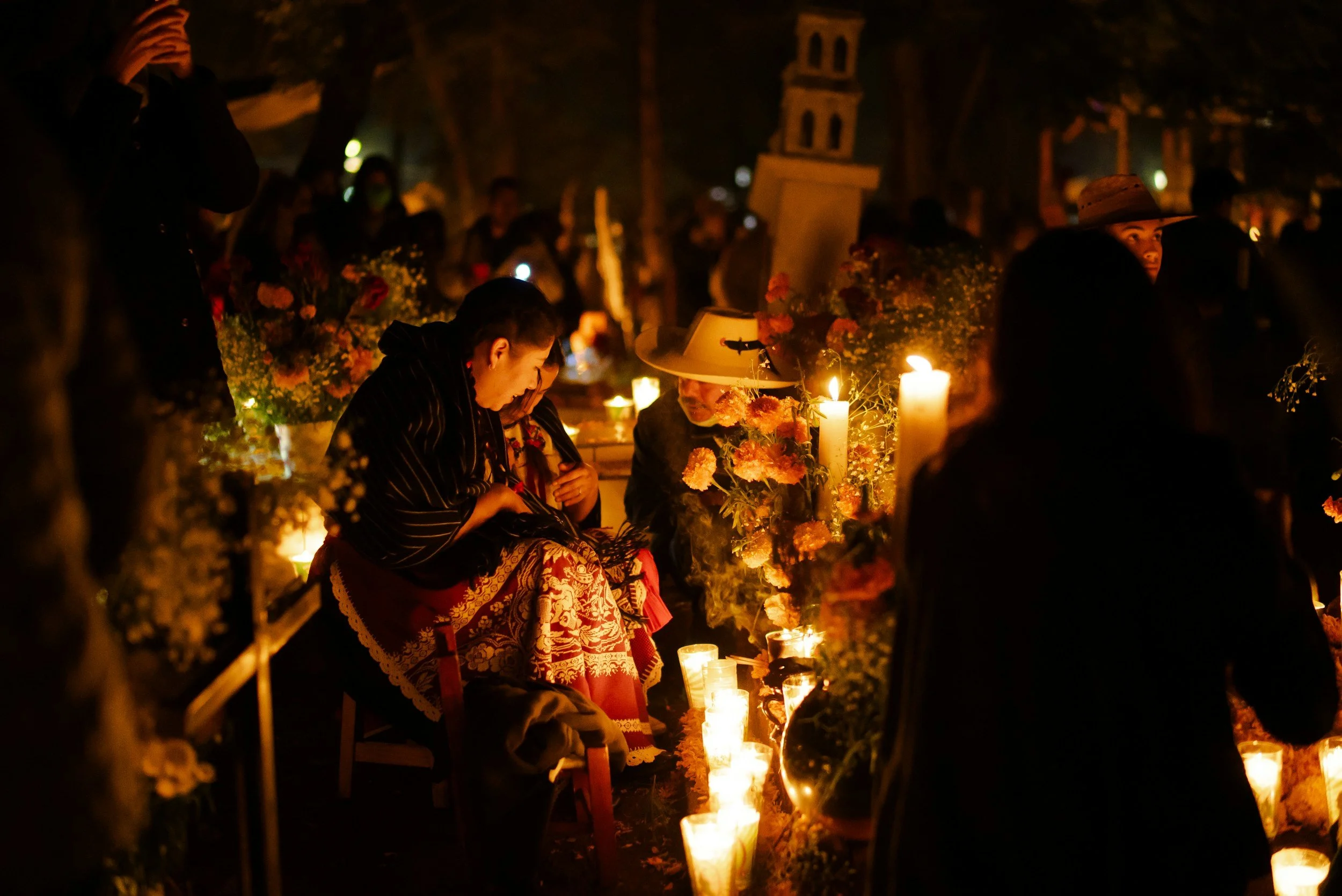 People gathered at a nighttime memorial or vigil, sitting among flowers and lit candles, with some wearing traditional Mexican clothing and sombreros.