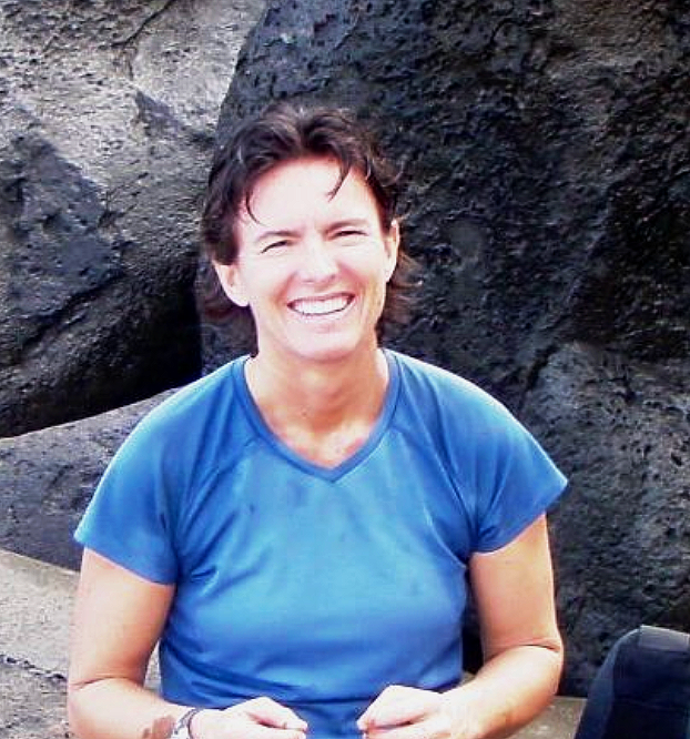 Woman with short brown hair in a blue shirt smiling in front of a rock outcrop.