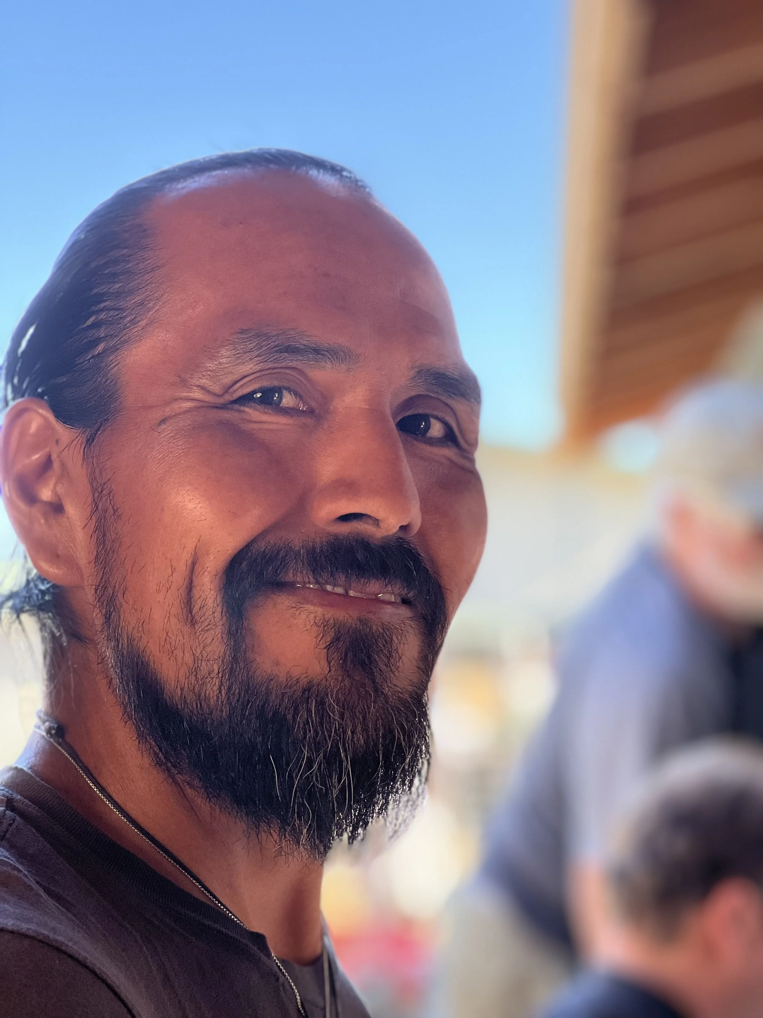 Close-up of a smiling man with a beard and mustache, wearing a dark shirt, against a blurred background with a blue sky and some wooden structure.