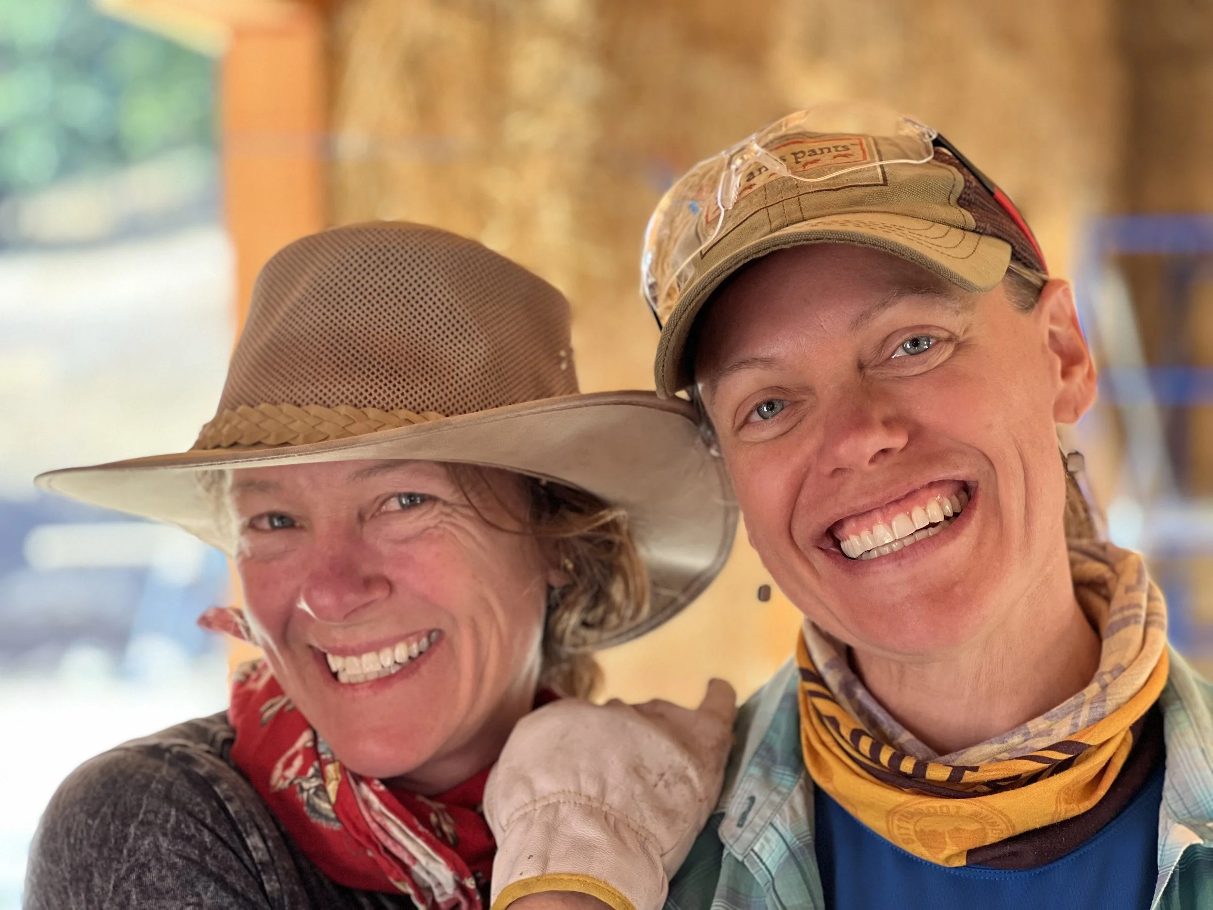 Two smiling women wearing hats and casual outdoor clothing, leaning close together against a colorful, blurred background.