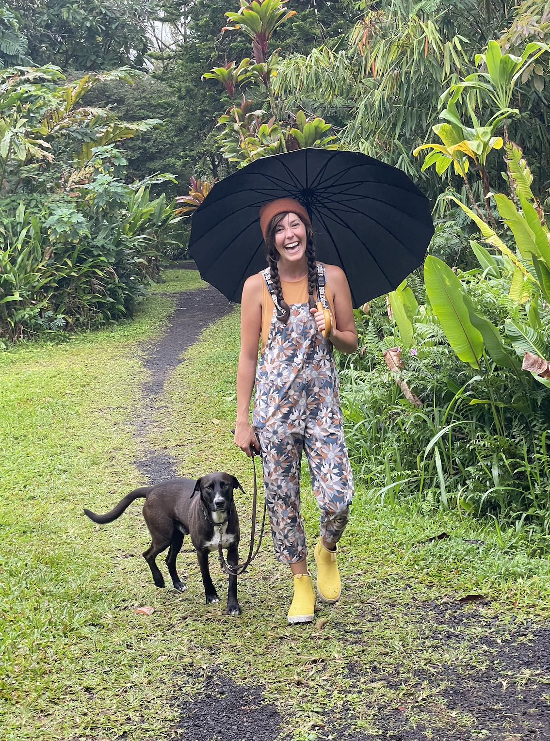 A woman with braided hair and a wide smile, wearing a brown beanie, a floral jumpsuit, yellow rain boots, and holding a black umbrella, walking on a muddy trail in a lush green jungle with a black dog on a leash.