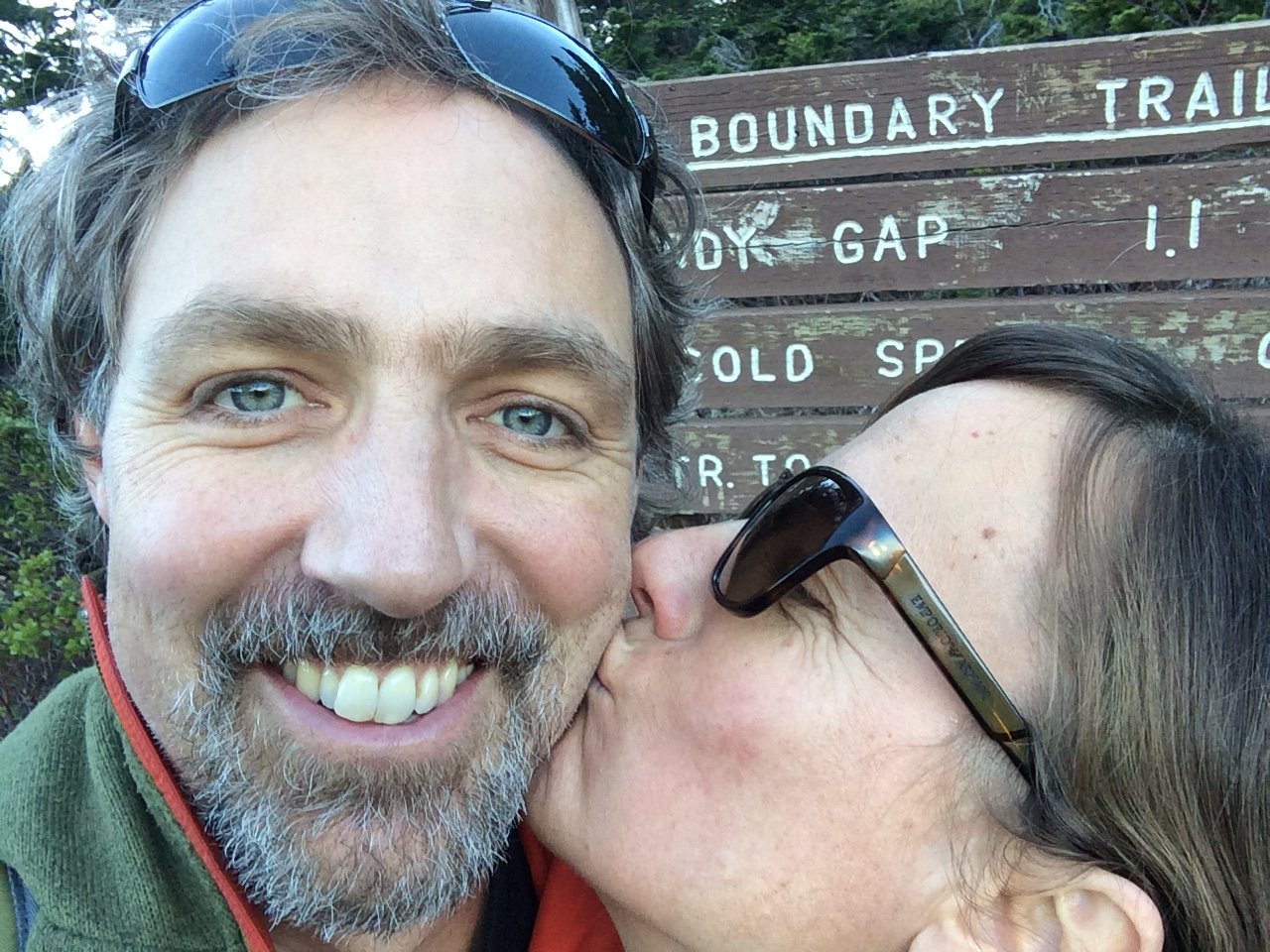 A man with gray hair and beard smiling while a woman wearing sunglasses kisses his cheek at a trailhead sign that reads 'Boundary Trail, Boy Gap 1.1, Old Spot' in a park or natural area.
