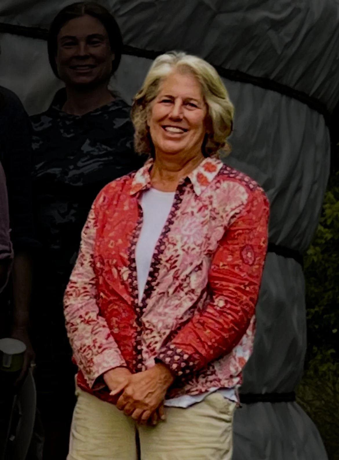Woman in a colorful red shirt and brown pants smiling with her hands crossed in front of her.