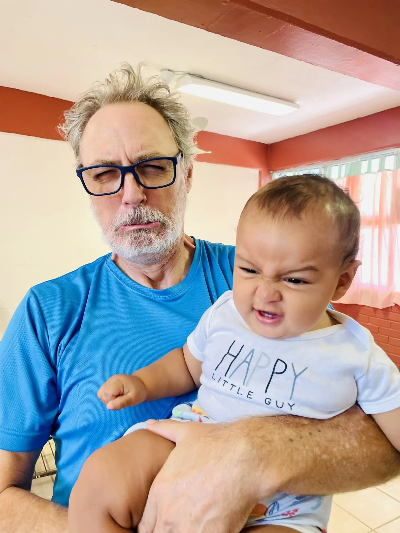 An older man with glasses and gray hair holding a young boy with a scrunched face. The boy is wearing a white T-shirt that says 'HAPPY LITTLE GUY.' The setting appears to be indoors, with a red and white wall and a window with curtains in the background.