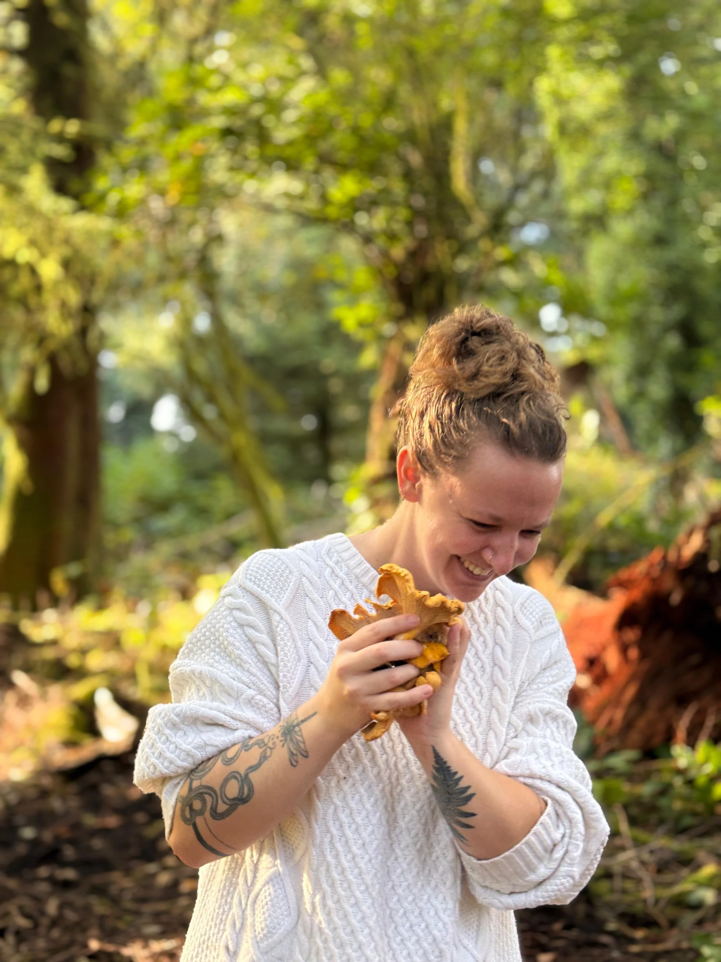 A woman with curly hair and tattoos on her arms, wearing a white knit sweater, is outdoors holding a large mushroom and smiling.