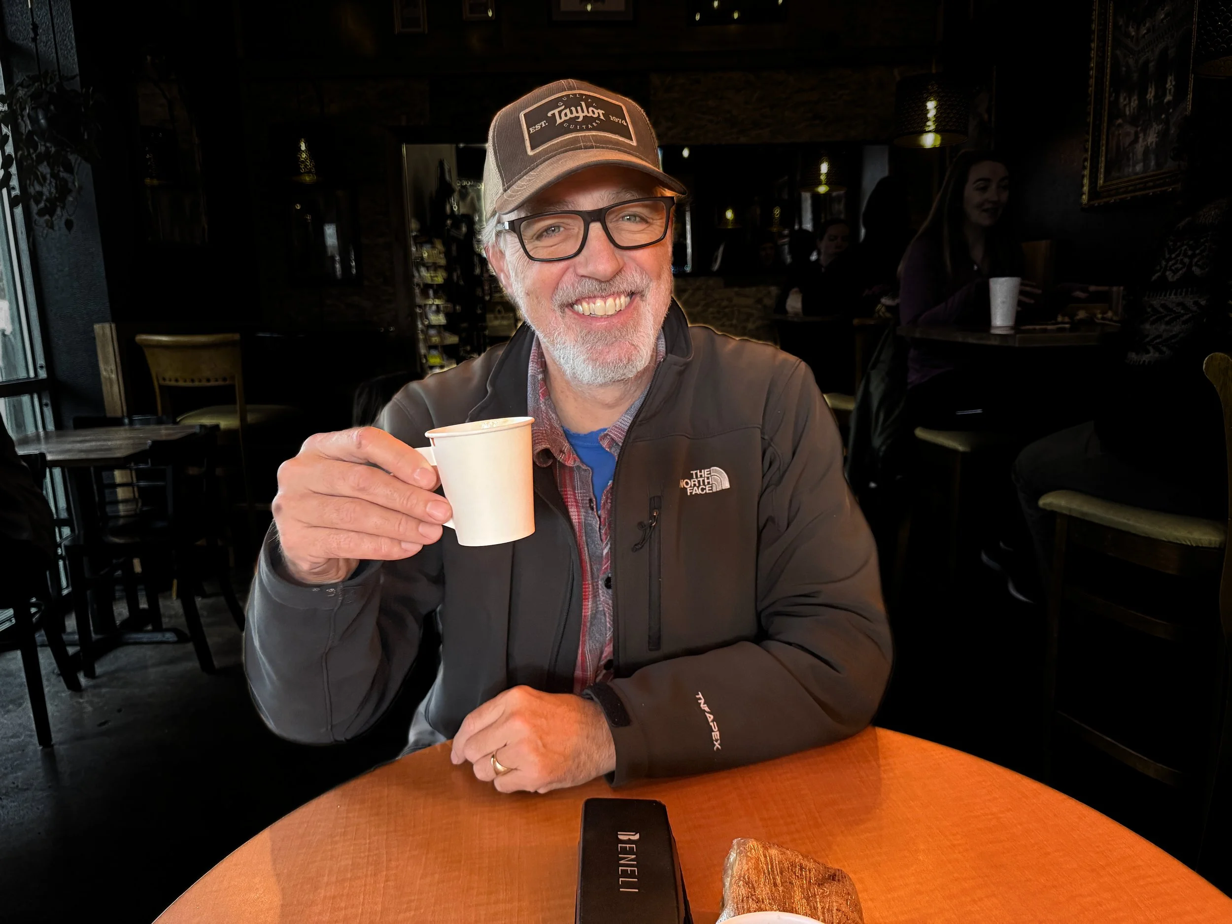 A smiling man with glasses and a baseball cap holding a small cup in a dimly lit indoor setting, with other people in the background.