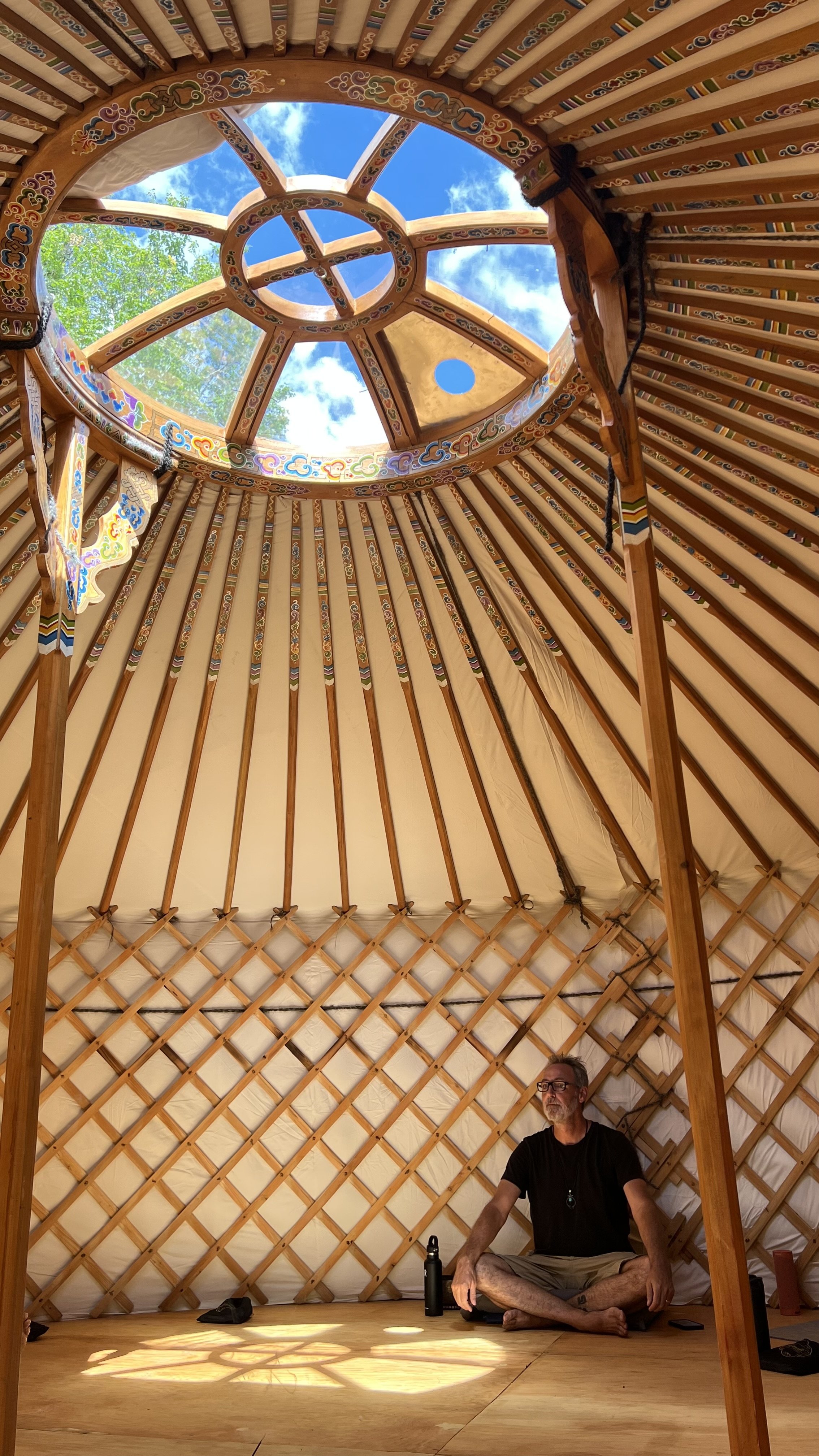 Interior of the yurt with a man wearing glasses and black shirt sitting cross-legged on wooden floor, sunlight filtering through the circular open roof, blue sky and some clouds visible.