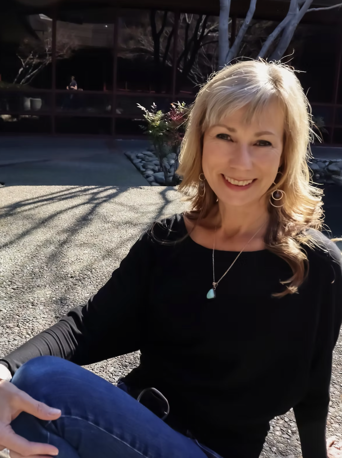 Blond woman in a black shirt and blue pants smiling and reclining casually.