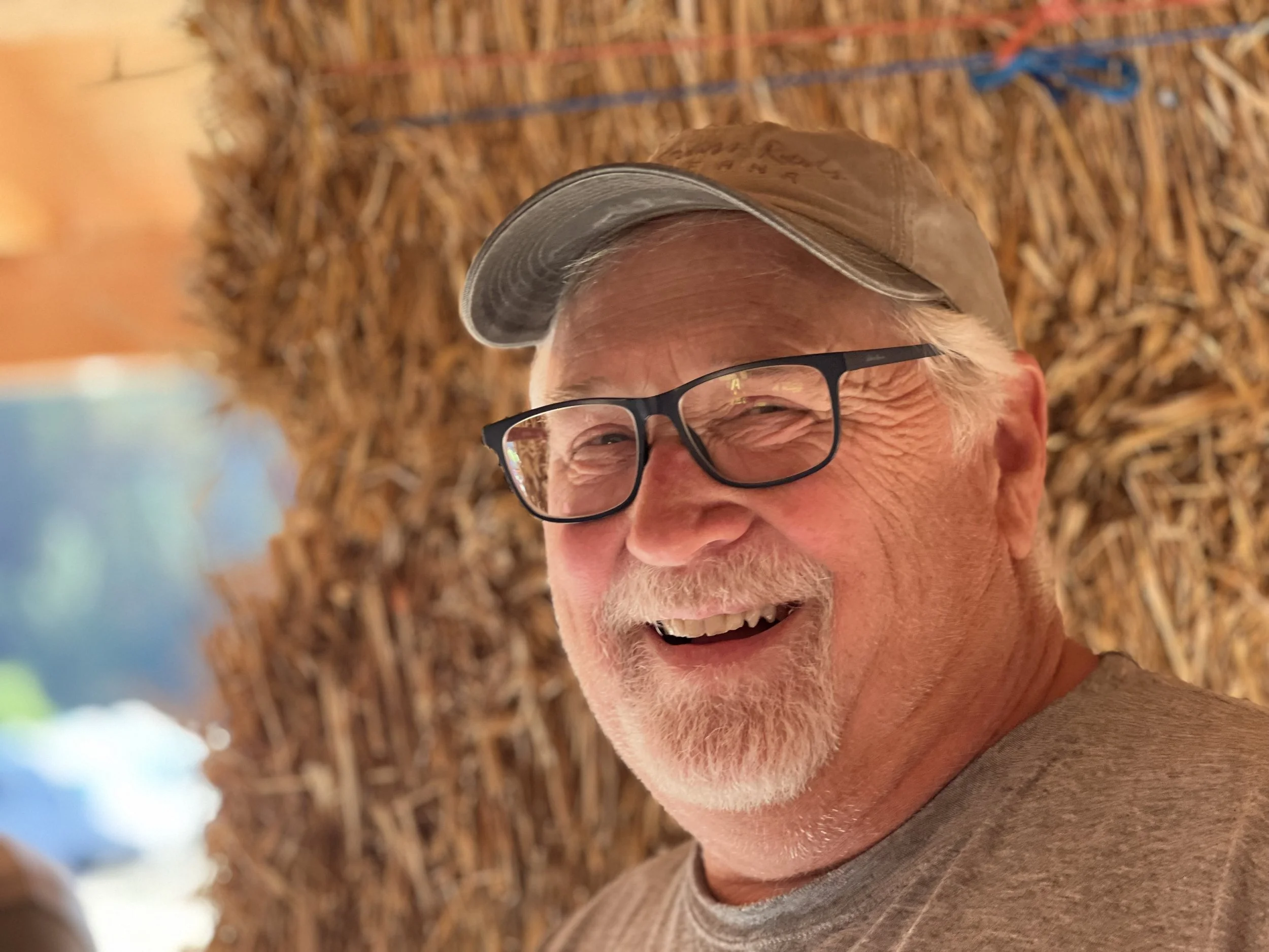 Close-up of smiling elderly man wearing glasses and a cap, with a background of straw or dried grass.
