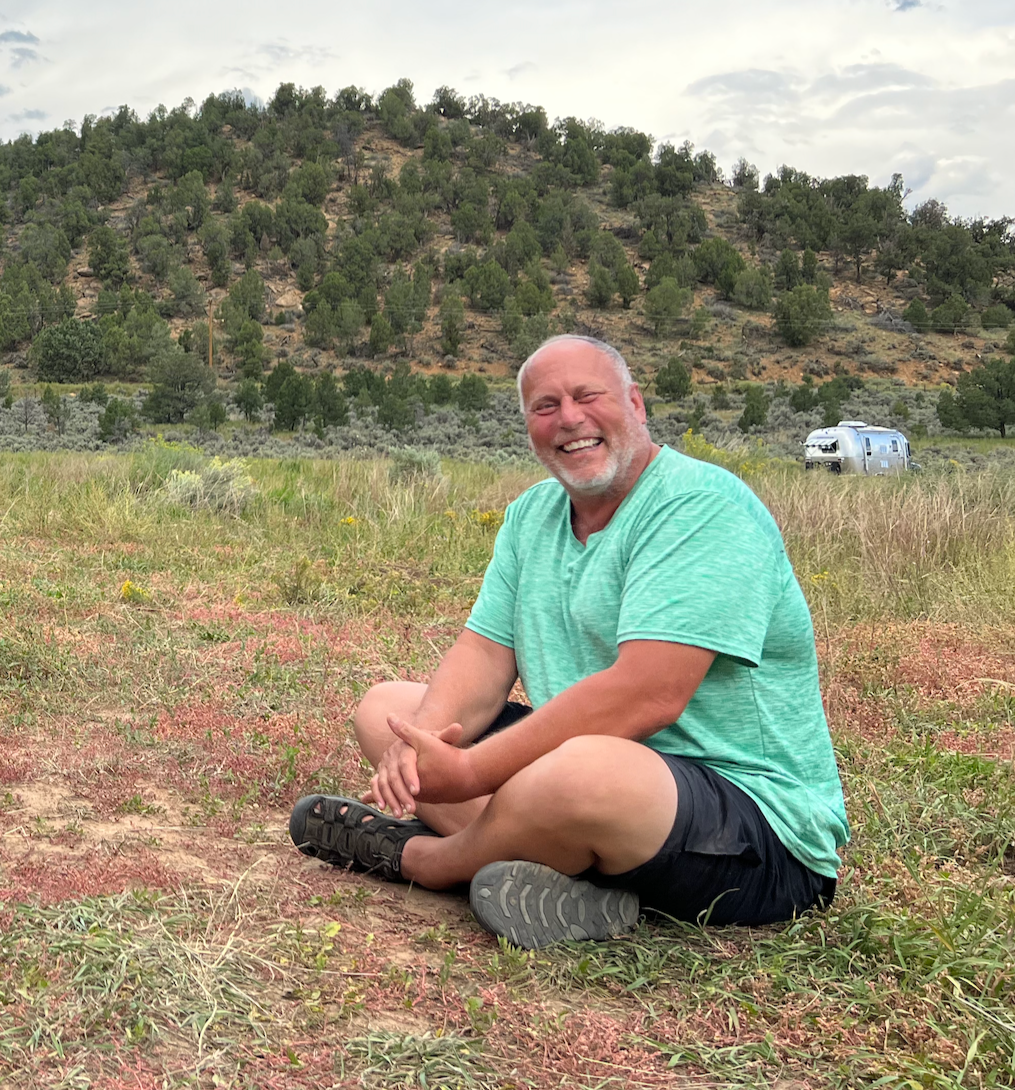 Older man with a big smile wearing a green shirt sitting in a field in front of a treed hill and a silver camping trailer.