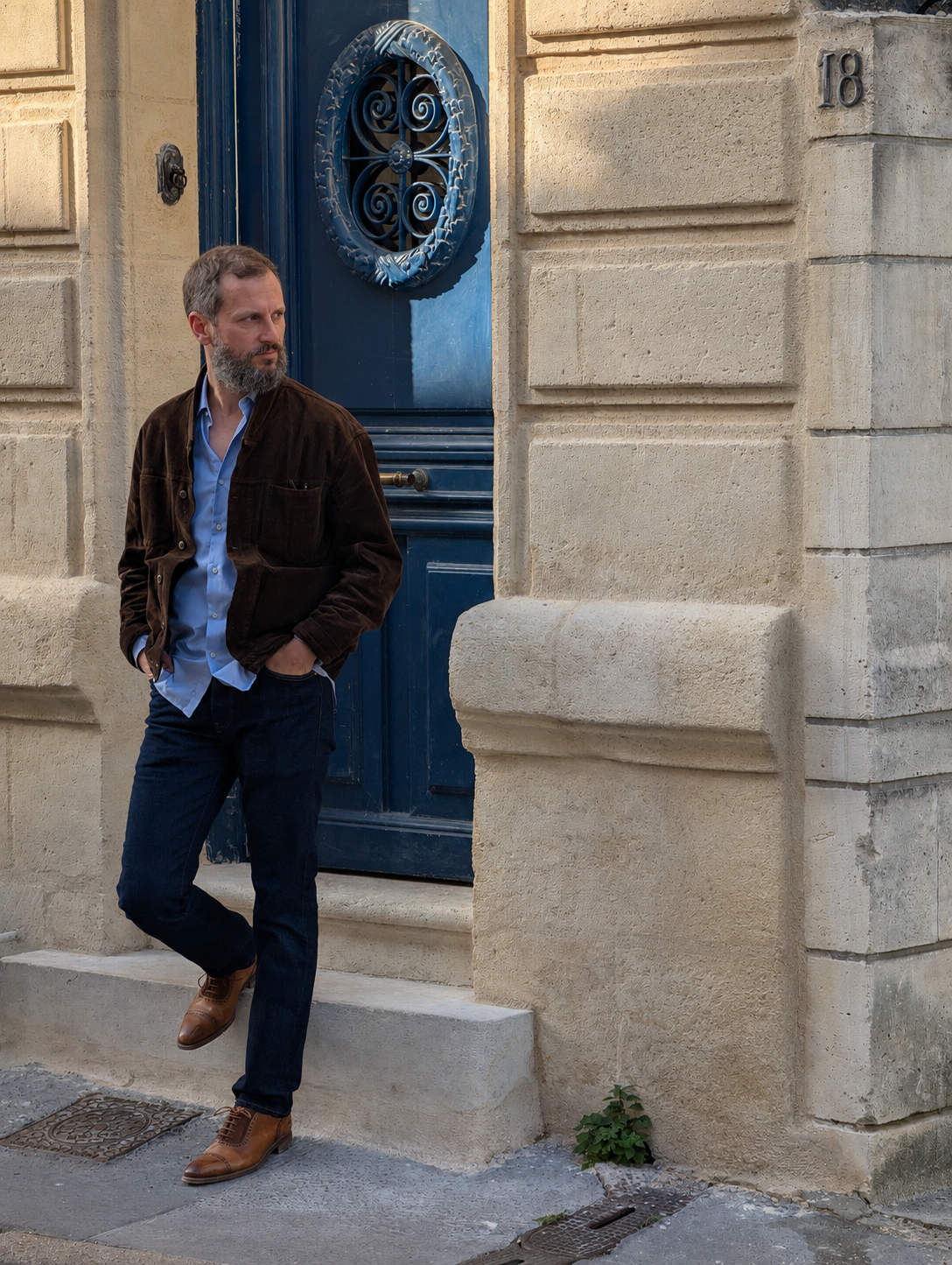 A man with a beard and brown hair leaning against a beige stone building with a blue door, standing on a sidewalk.
