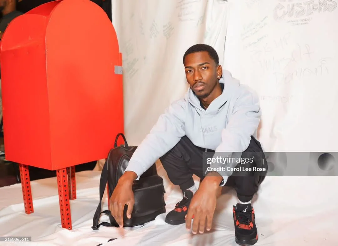 A young man squatting on the floor next to a black backpack and a bright red mailbox or donation box. He is wearing a light gray hoodie, dark shorts, and black and red sneakers, with a serious expression on his face. The background is white with faint writing or drawings.