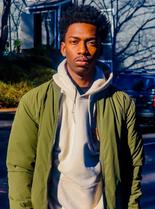 Young man with curly hair wearing a green jacket over a light gray hoodie, standing outdoors with trees, a building, and cars in the background.