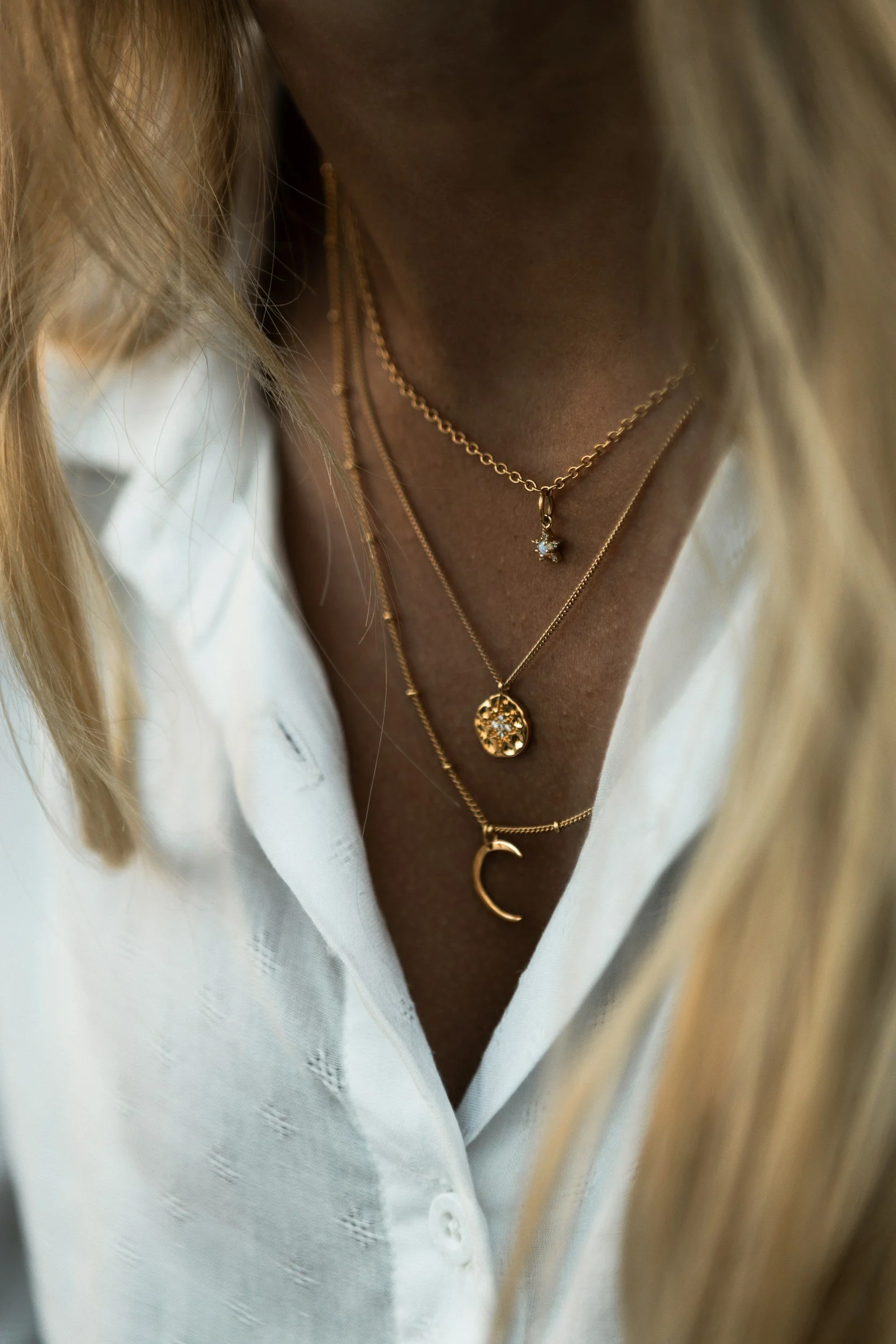Close-up of a person's neck and chest wearing layered gold necklaces with different pendants and a white button-up shirt.