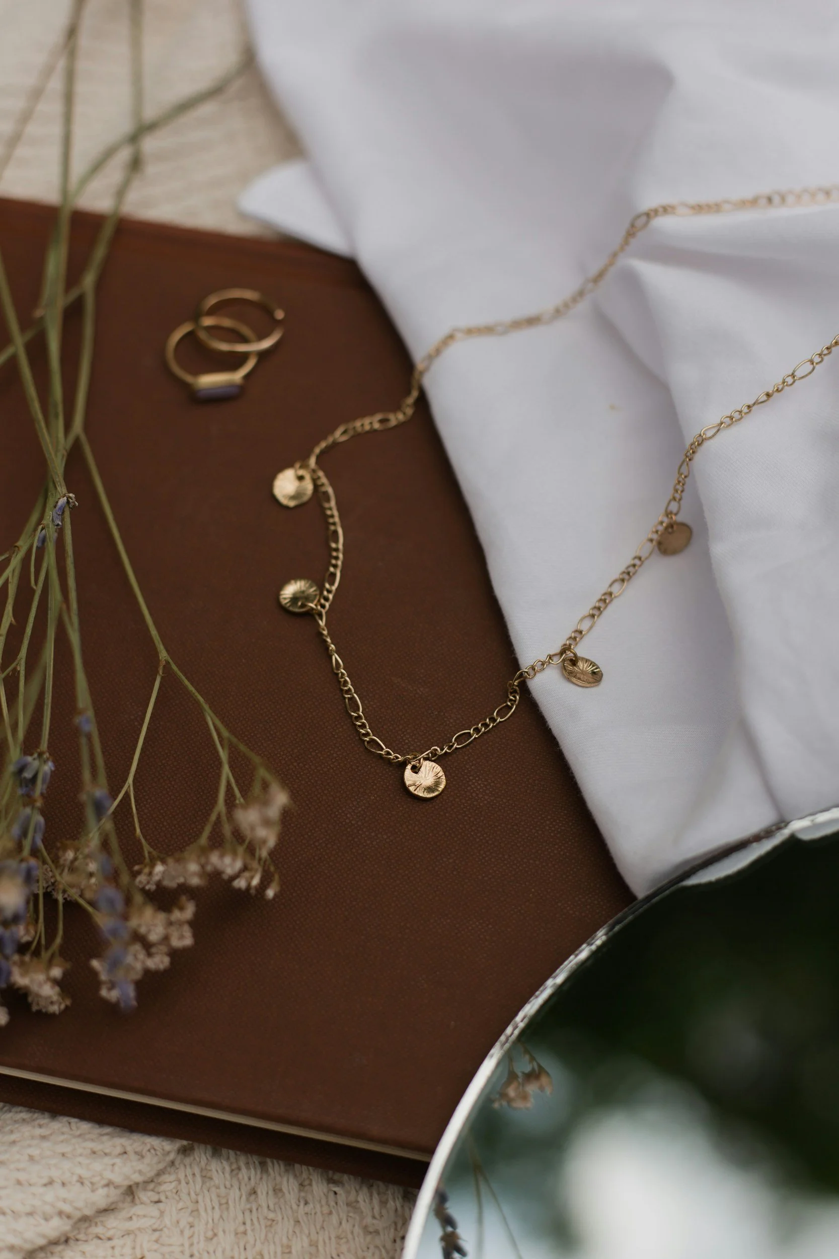A gold chain necklace with small round charms, a pair of gold hoop earrings, dried lavender sprigs, a white fabric item, a wooden surface, and a green ceramic vessel.